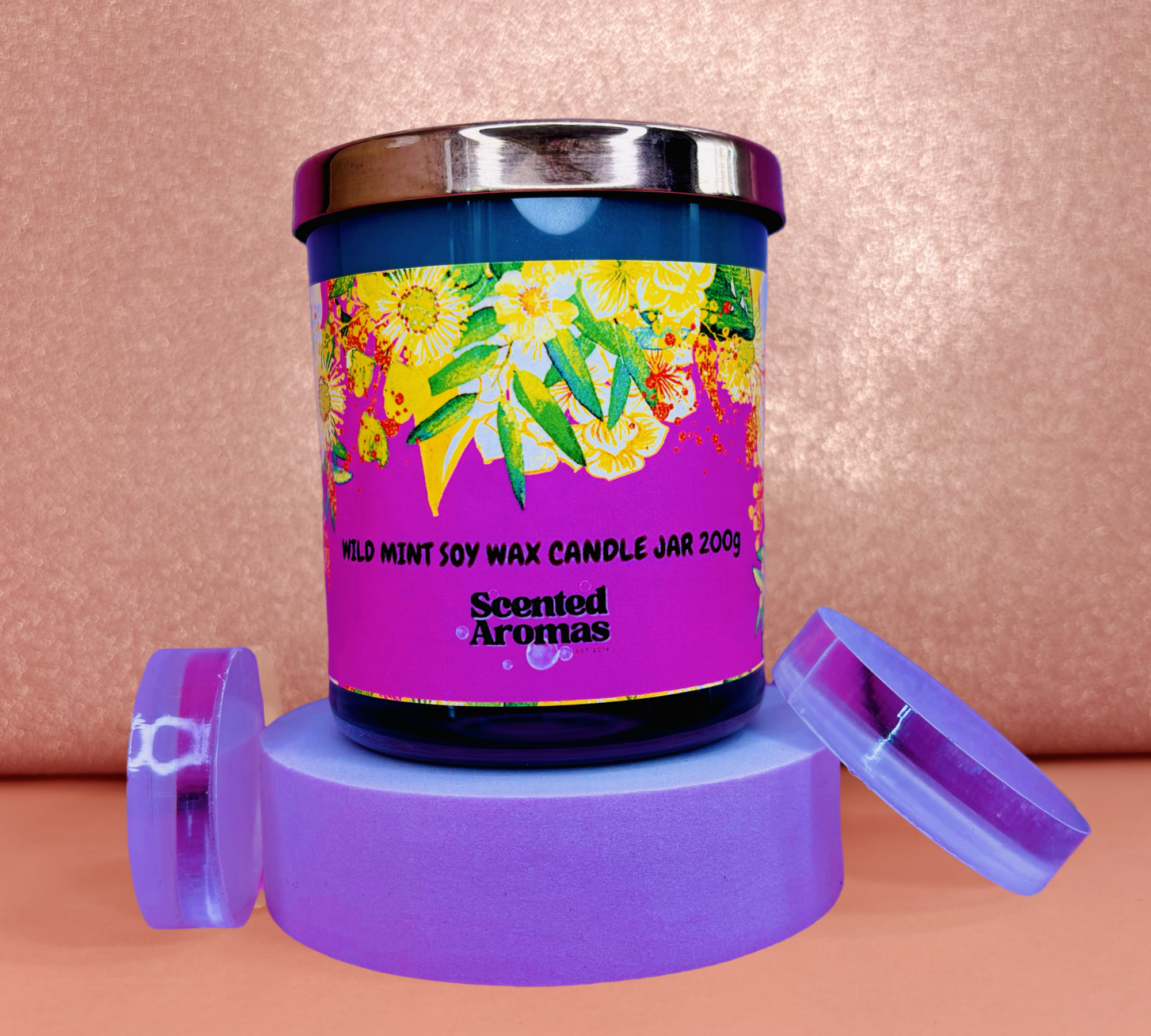 The "Wild Mint Candle - 200g" by The Soap Gal x, boasting a refreshing aroma, sits elegantly on a purple stand. This soy wax candle jar, embellished with a vibrant floral design, captivates your senses against its charming pink backdrop as the lid rests gracefully beside it.