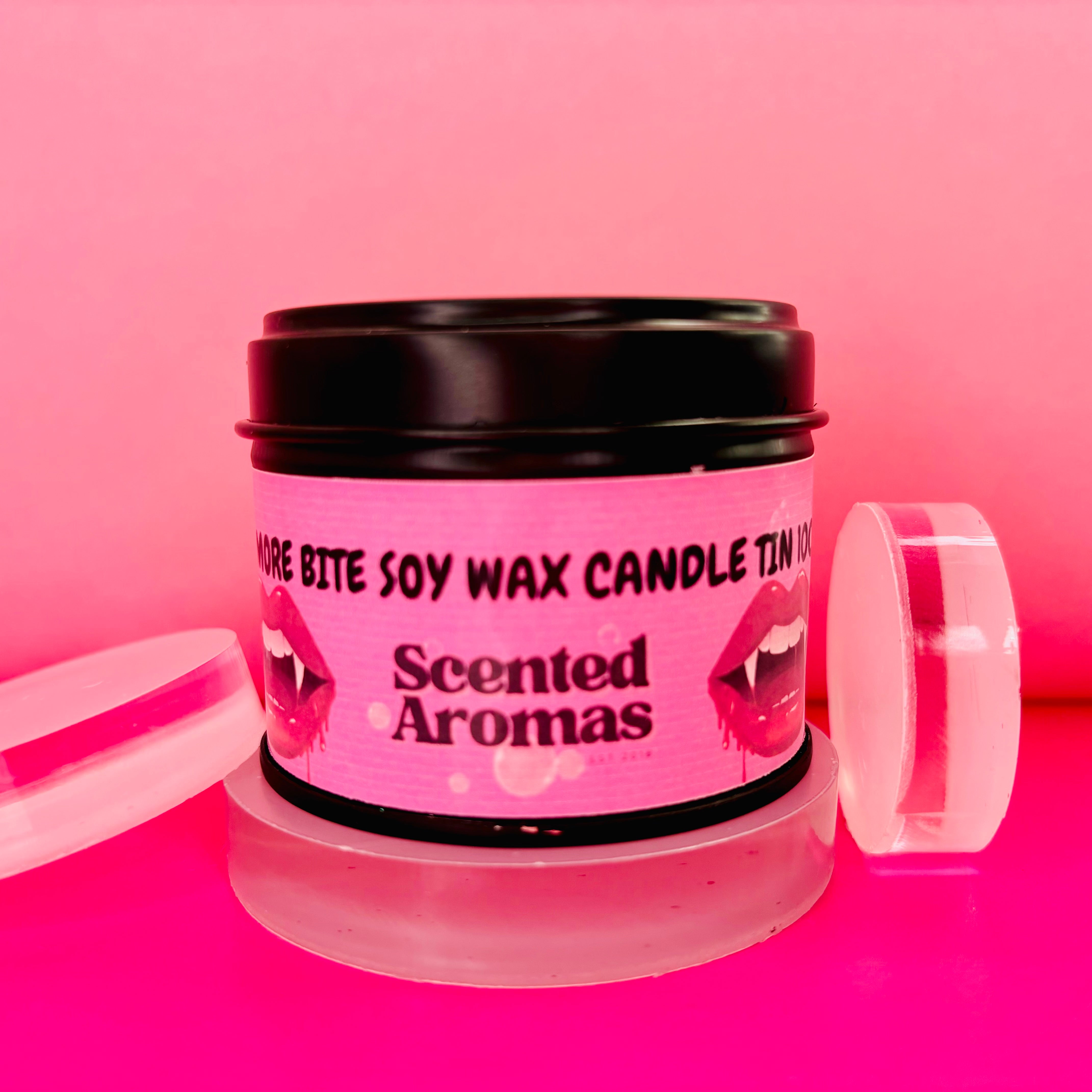 A pink tin labeled with "The Soap Gal x" features an illustration of lips and promises long-lasting fragrances. The text reads "One More Bite Candle Tin - 100ml," against a matching pink background.