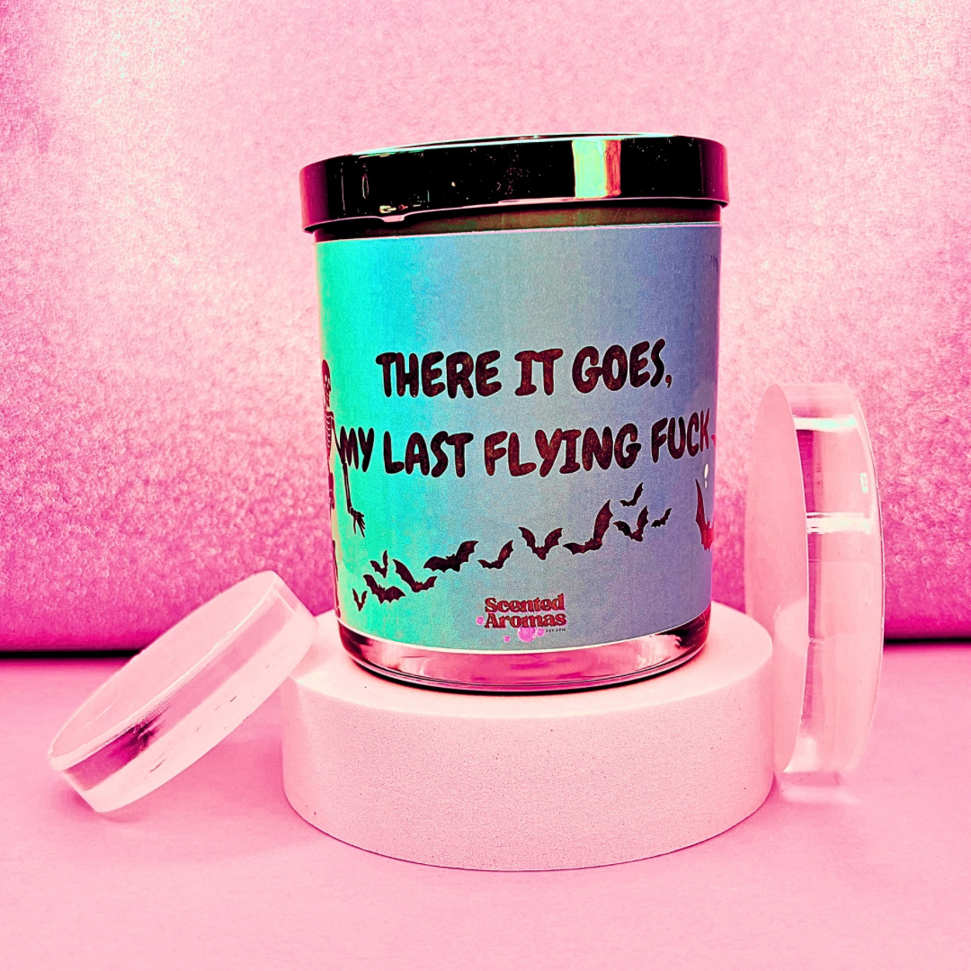 A soy wax candle with a lid sits on a pink background. The label humorously reads, "There it goes, my last flying care," adorned with bird silhouettes and the brand name "The Soap Gal x." This funny quote candle makes for a perfect gift.