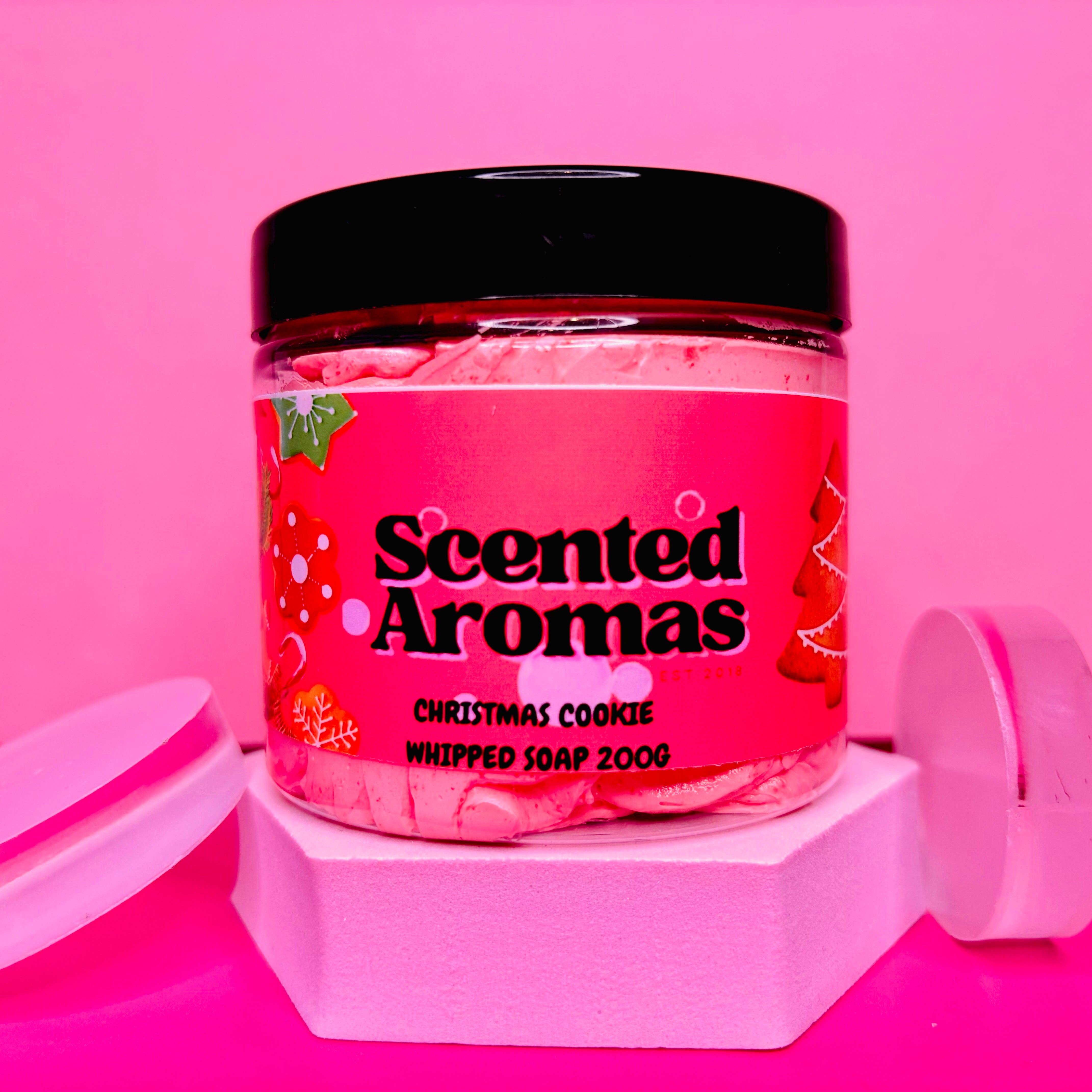 A jar of The Soap Gal x's Christmas Cookies Whipped Soap Shave Butter, 200g, featuring a sleek black lid, sits elegantly against a pink background. Perfect for indulging your senses.