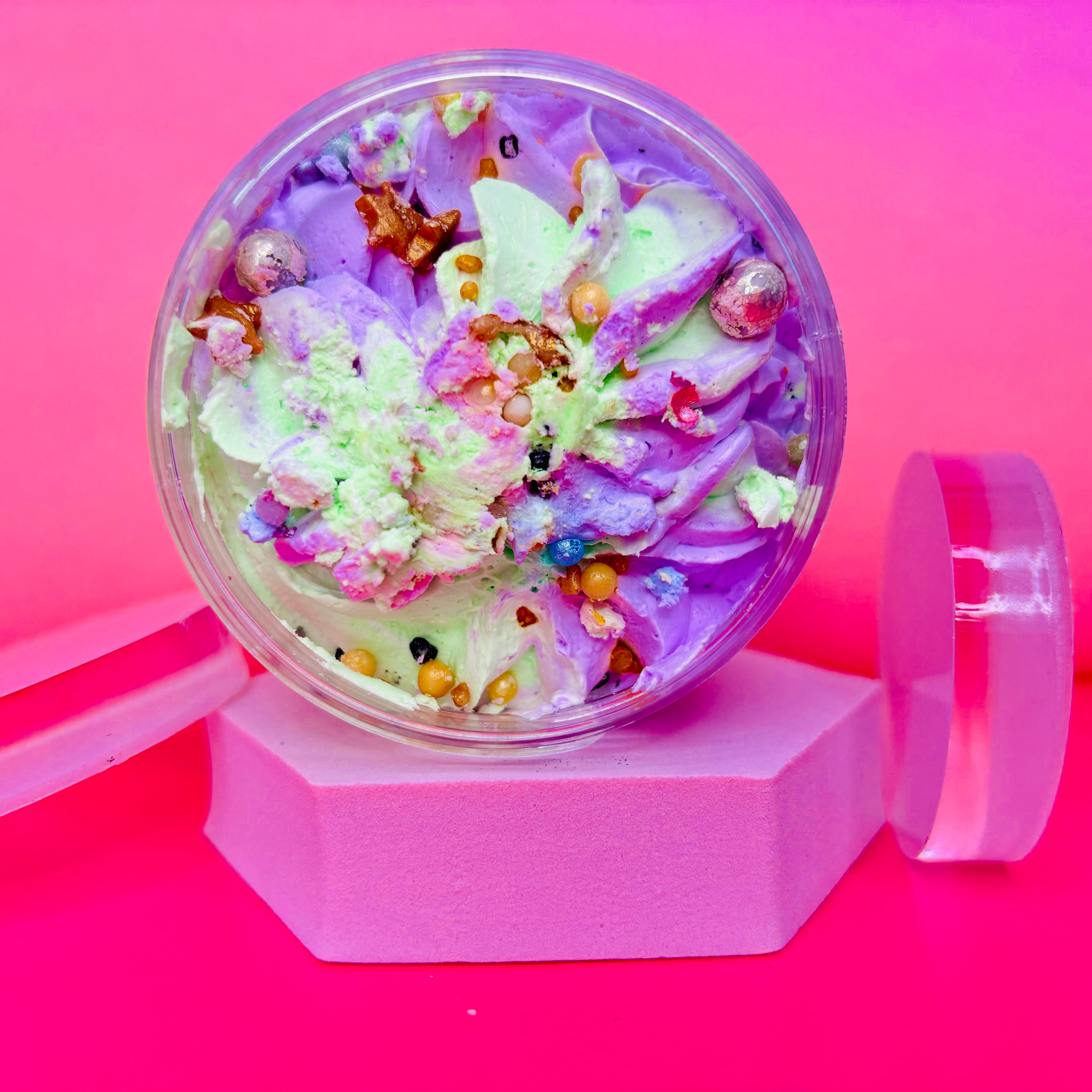 A round container of Scented Aromas' Alien Encounter Whipped Soap Shave Butter 200g with sprinkles sits on a pink hexagonal stand against a bright pink backdrop.