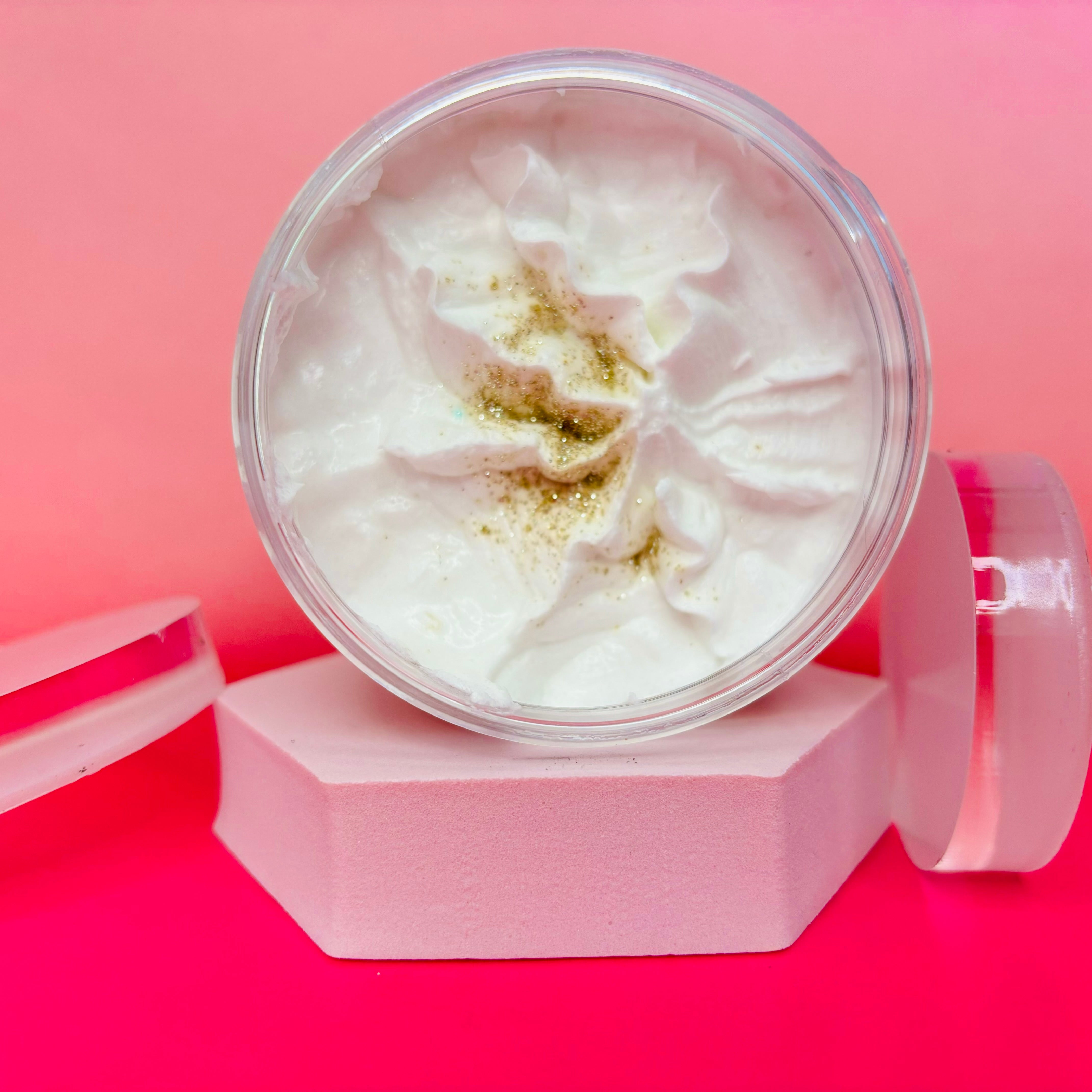 A jar of Almond Milk and Honey Sugar Body Scrub by Scented Aromas, topped with gold sprinkles, rests elegantly on a pink hexagonal stand against a pink and red backdrop. Perfect for smooth, glowing skin.