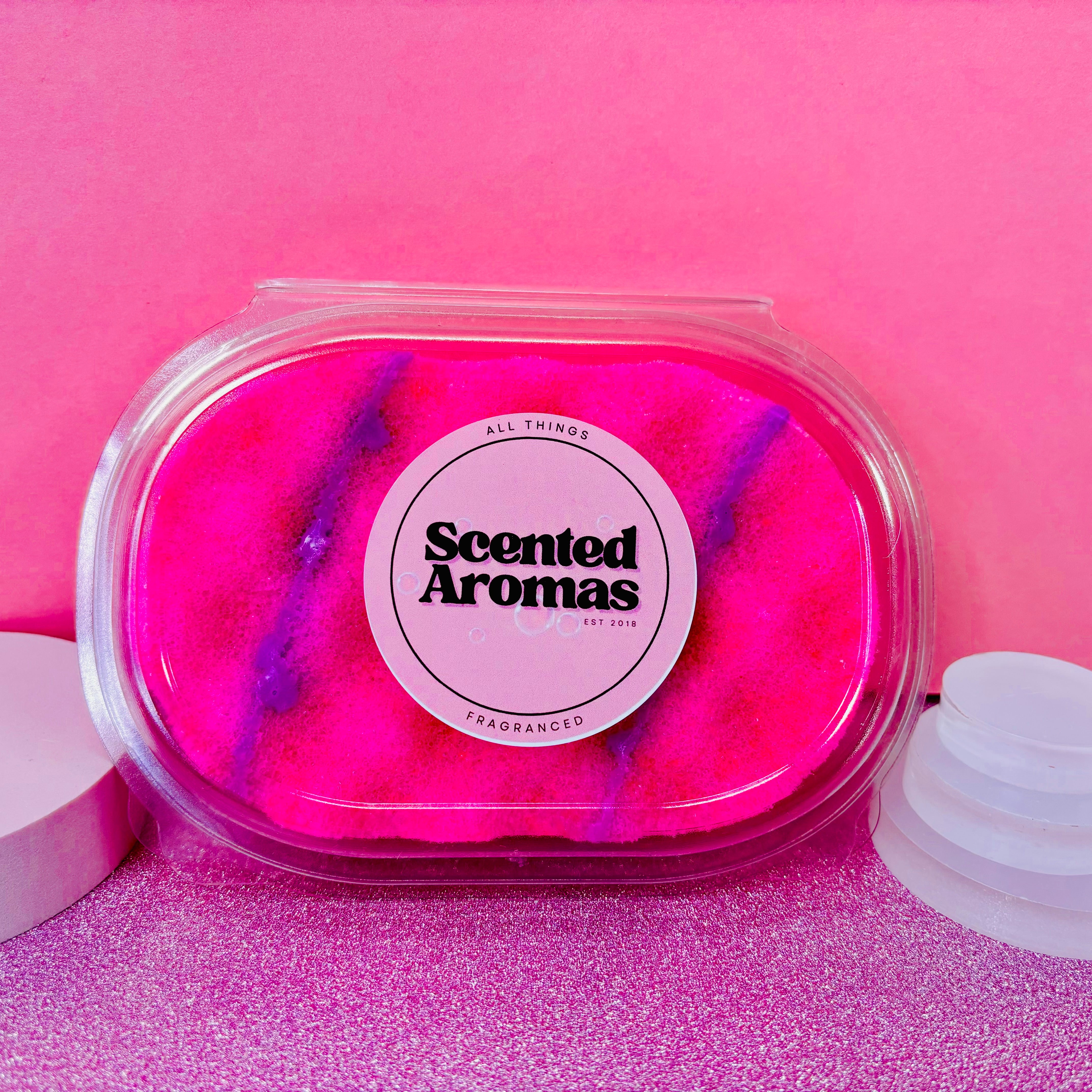 An oval container filled with vibrant pink and purple sand, labeled "Scented Aromas," evokes the luxury of a Millionairess Perfume Soap Sponge by The Soap Gal x, set against a pink background.