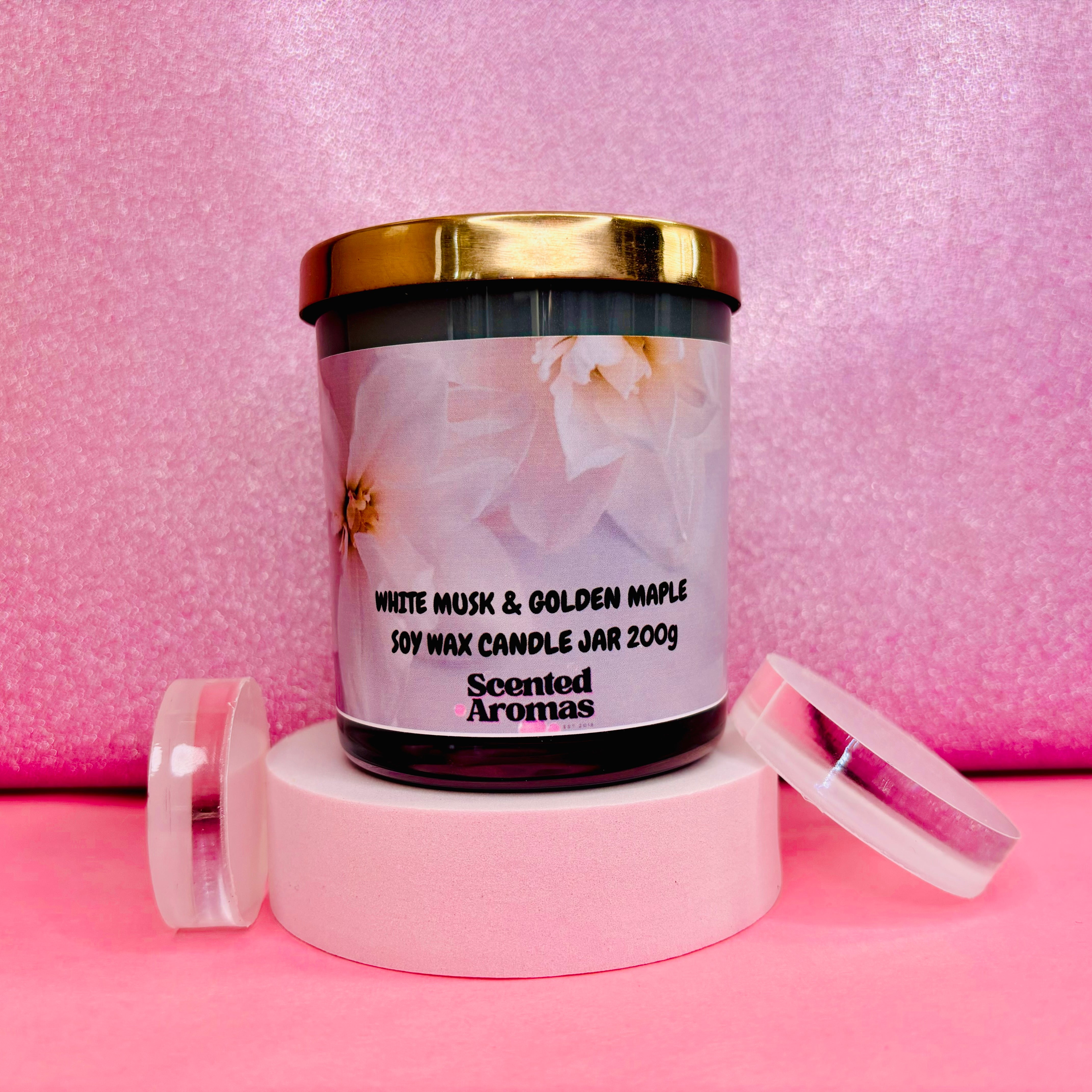 A jar candle labeled "White Musk and Maple Glass Candle 200g" by The Soap Gal x rests elegantly on a round platform against a pink background, evoking the style of a Mrs. Hinch inspired candle collection.