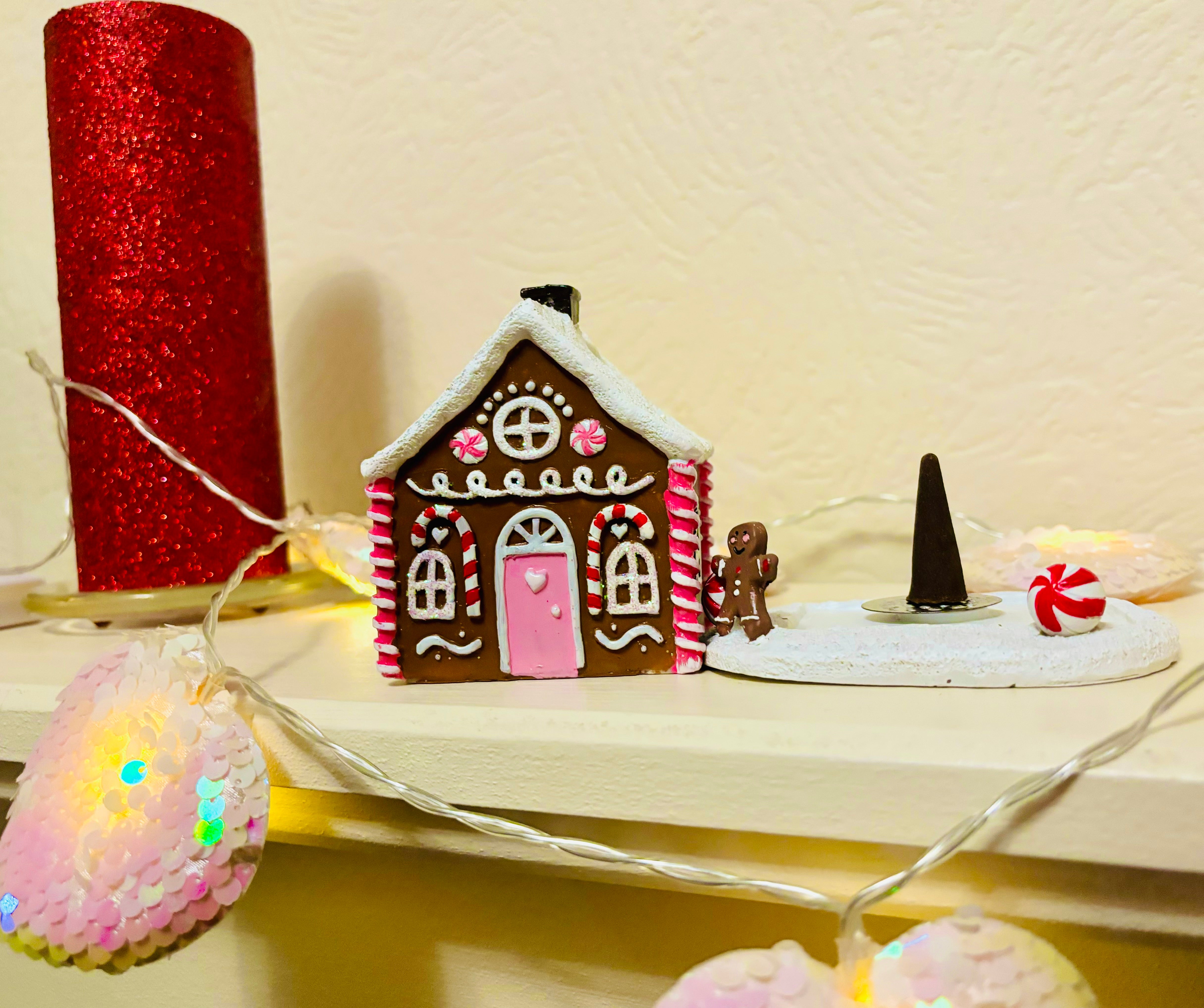 The Soap Gal x presents their Pink Gingerbread House Incense Burner, adorned with a candy cane design and featuring a whimsical small figure in a witch hat. Enhance your enchanting festive decor with the added charm of a red glitter candle and glowing string lights, creating the perfect Christmas fairytale ambiance.