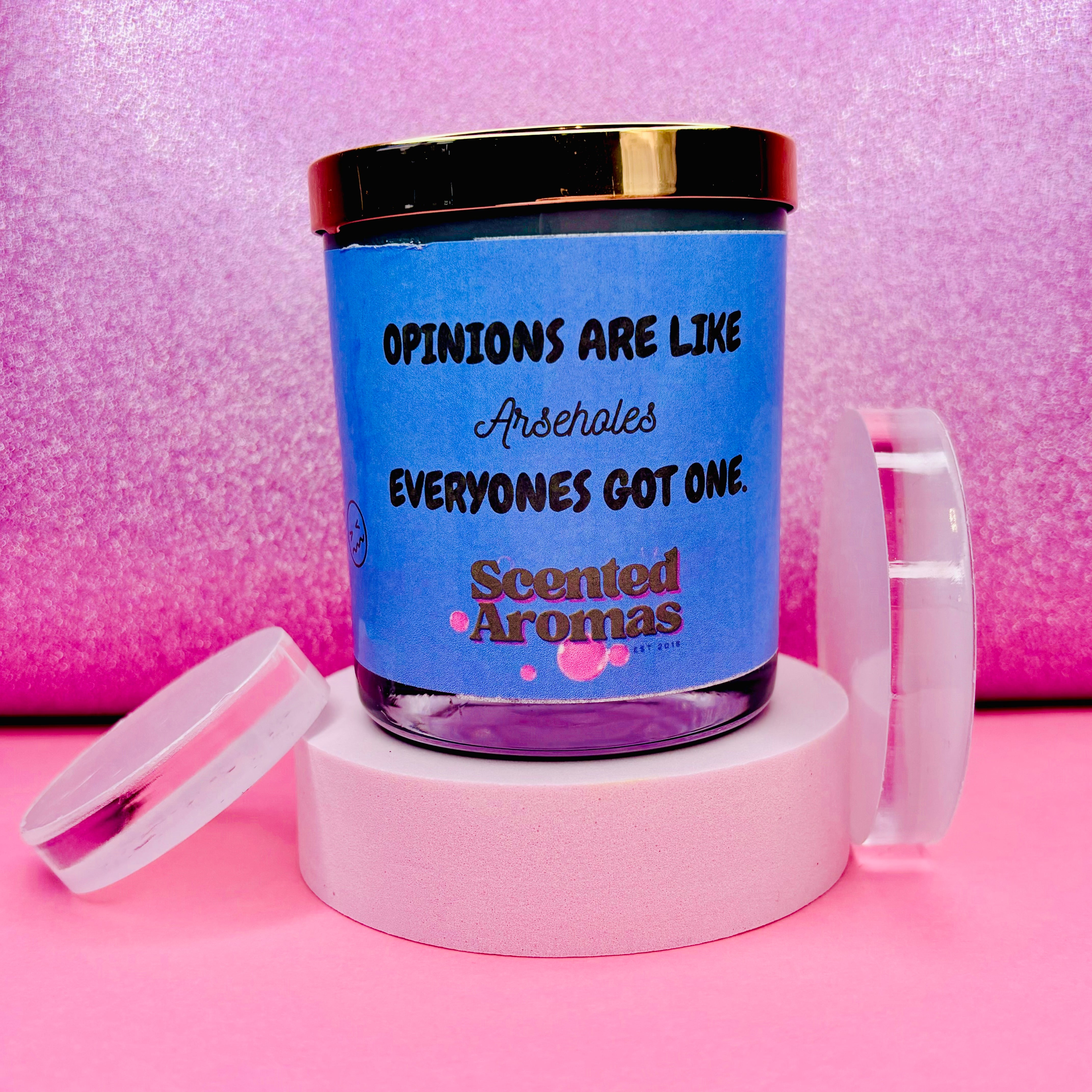 The Oh So Opinionated Funny Quote Candle by The Soap Gal x, featuring a blue label with the phrase "Opinions are like arseholes, everyone's got one," sits on a pink background with its lid removed. Made from 100% pure soy wax, this 200g candle is an amusing gift choice.