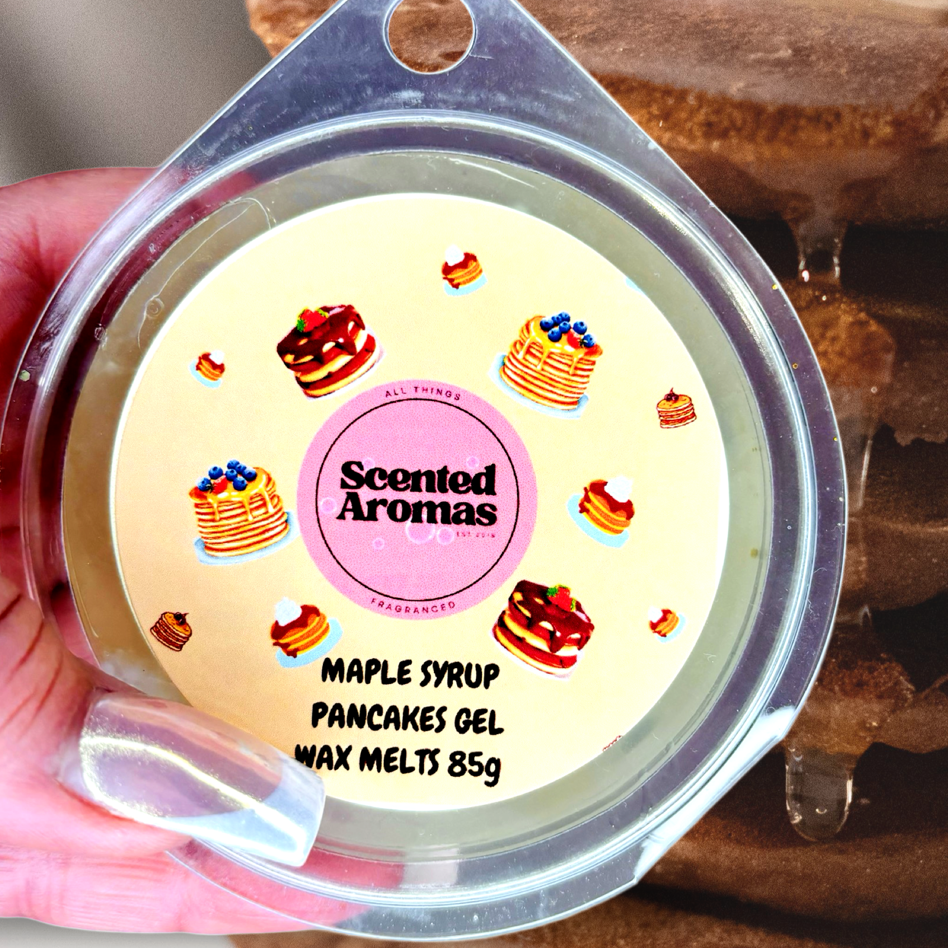 A hand holds a container of The Soap Gal x Maple Syrup Pancakes Gel Wax Melts, 85g. The label features delicious maple syrup pancakes, emphasizing the enticing fragrance inside.