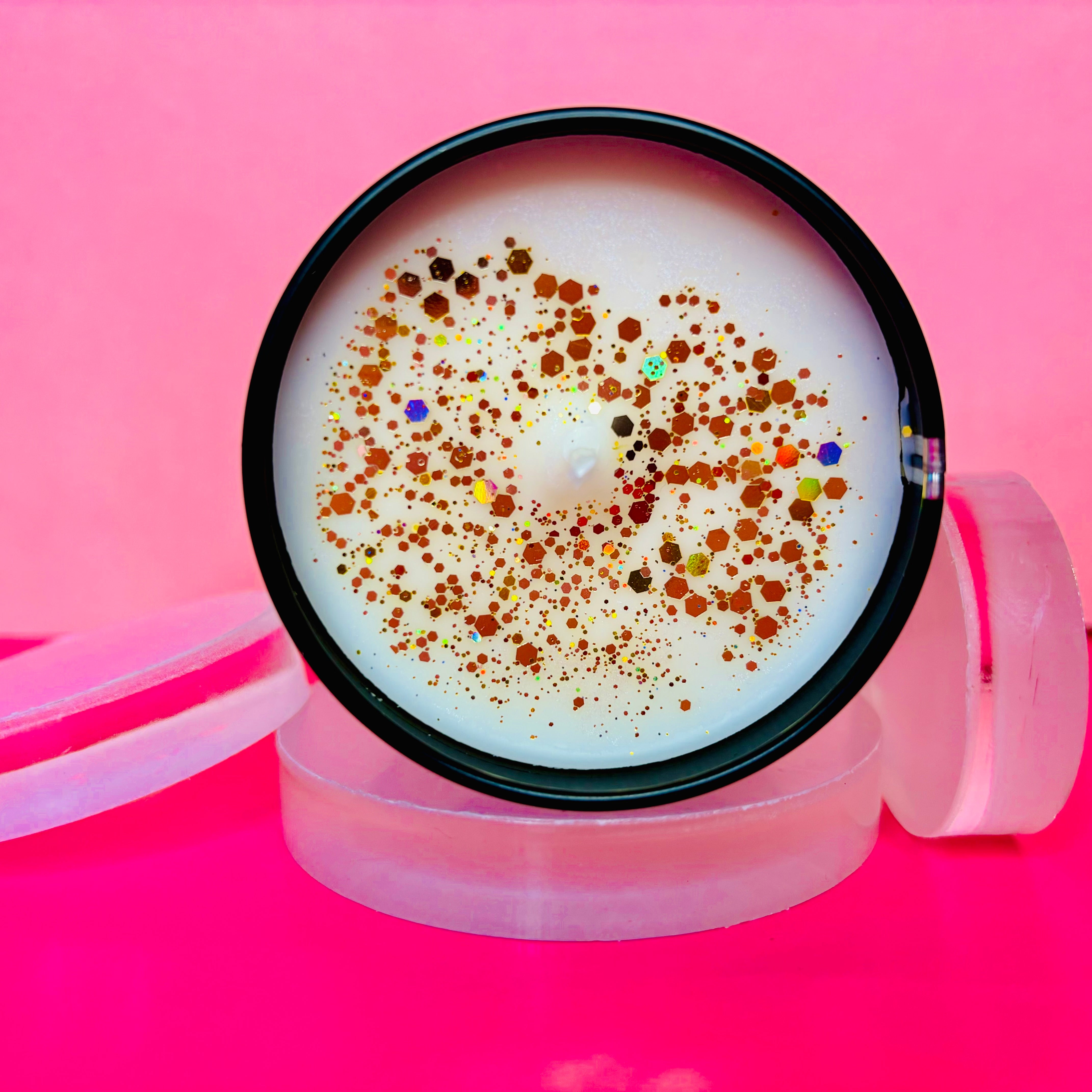 A round tin from The Soap Gal x, filled with their Salted Caramel Candle in a 100ml size, is adorned with golden glitter and rests gracefully on a series of clear discs against a pink background, capturing an essence of sophistication.