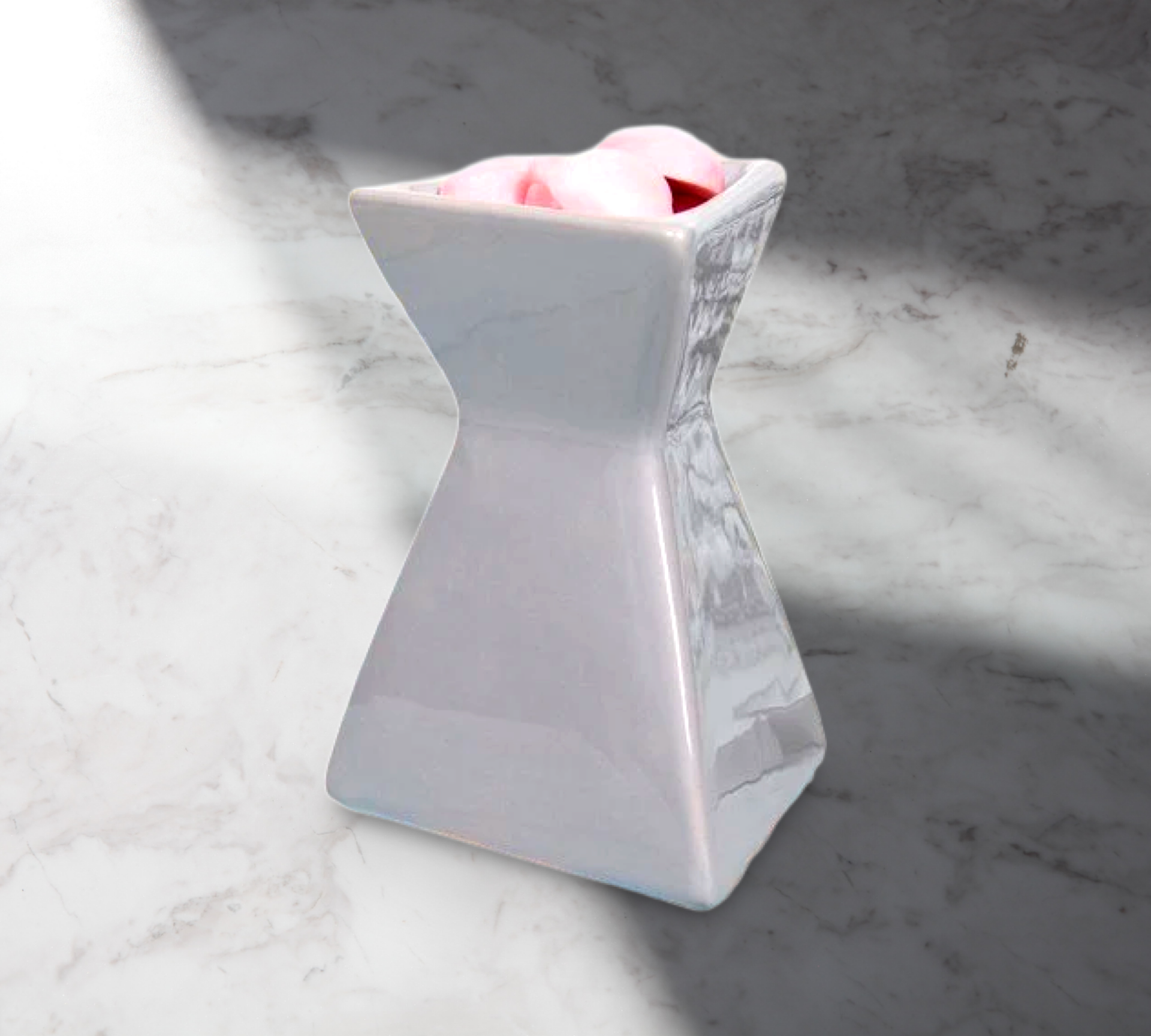 A white, angular vase adorned with pink flowers sits elegantly on a marble surface, partly obscured by shadows. Close by, "The Soap Gal x Grey Triangle Wax Warmer" gently disperses delightful home fragrances, crafting an inviting ambience.