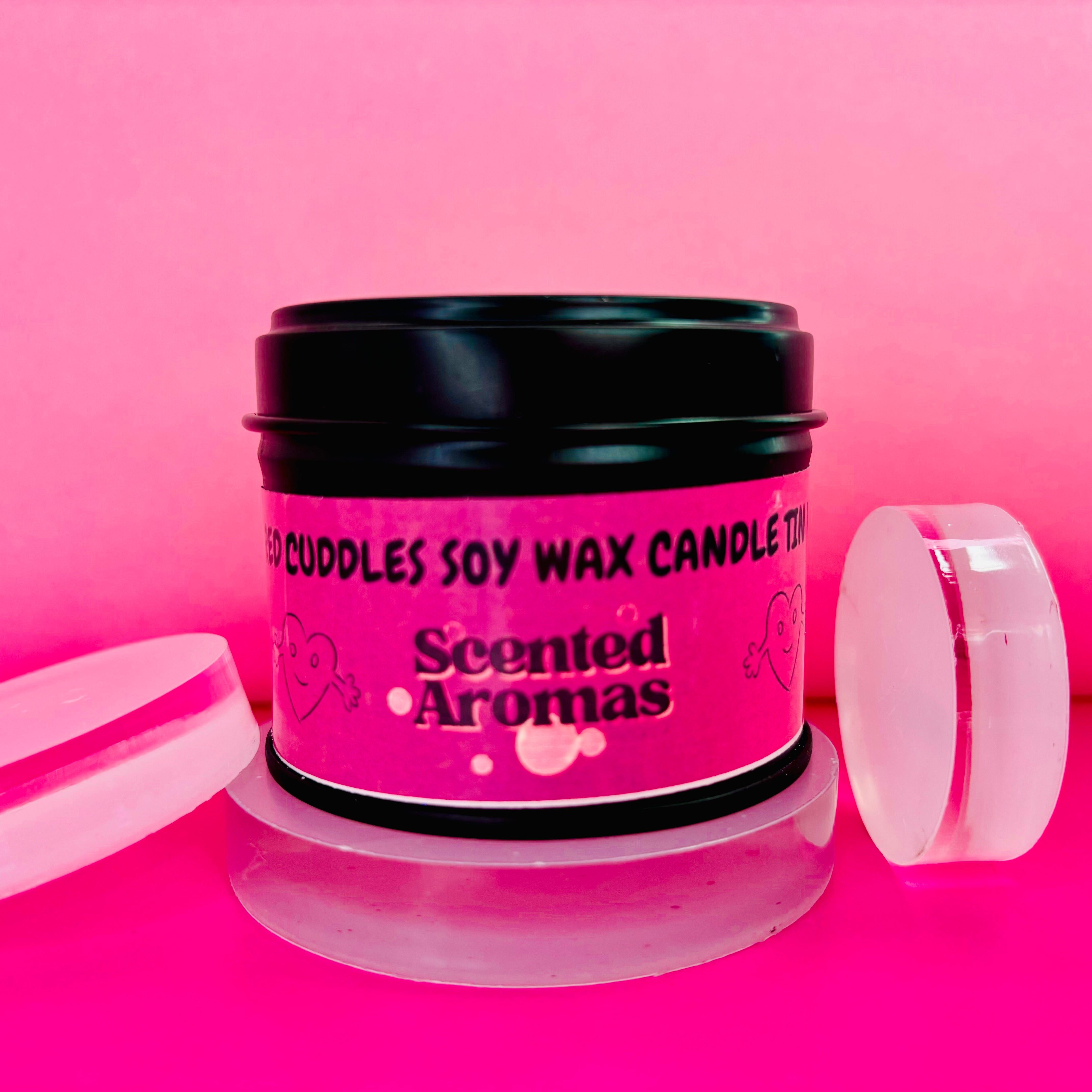 A black tin candle from The Soap Gal x, labeled "Sugared Cuddles Candle Tin - 100ml," rests on a clear, circular stand against a pink backdrop. It features a pink sticker adorned with hearts and is crafted from soy wax to provide a long-lasting fragrance that delights the senses.