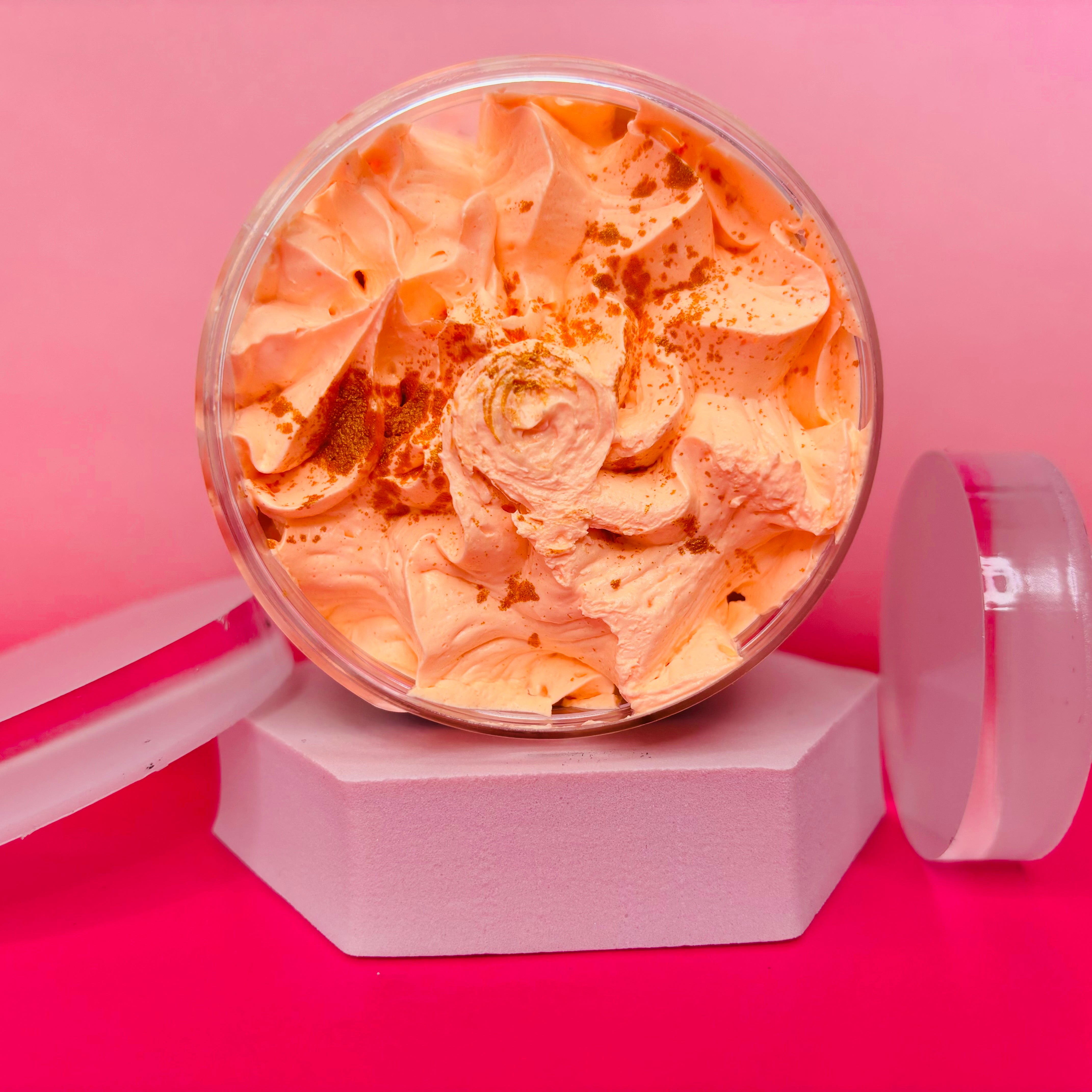 A round container of The Soap Gal x's Brazilian Bum Cream Sugar Body Scrub 200g, with its swirled orange cream, sits on a white hexagonal stand against a pink backdrop. Its transparent lid lies beside it—ideal for elevating your skincare routine.