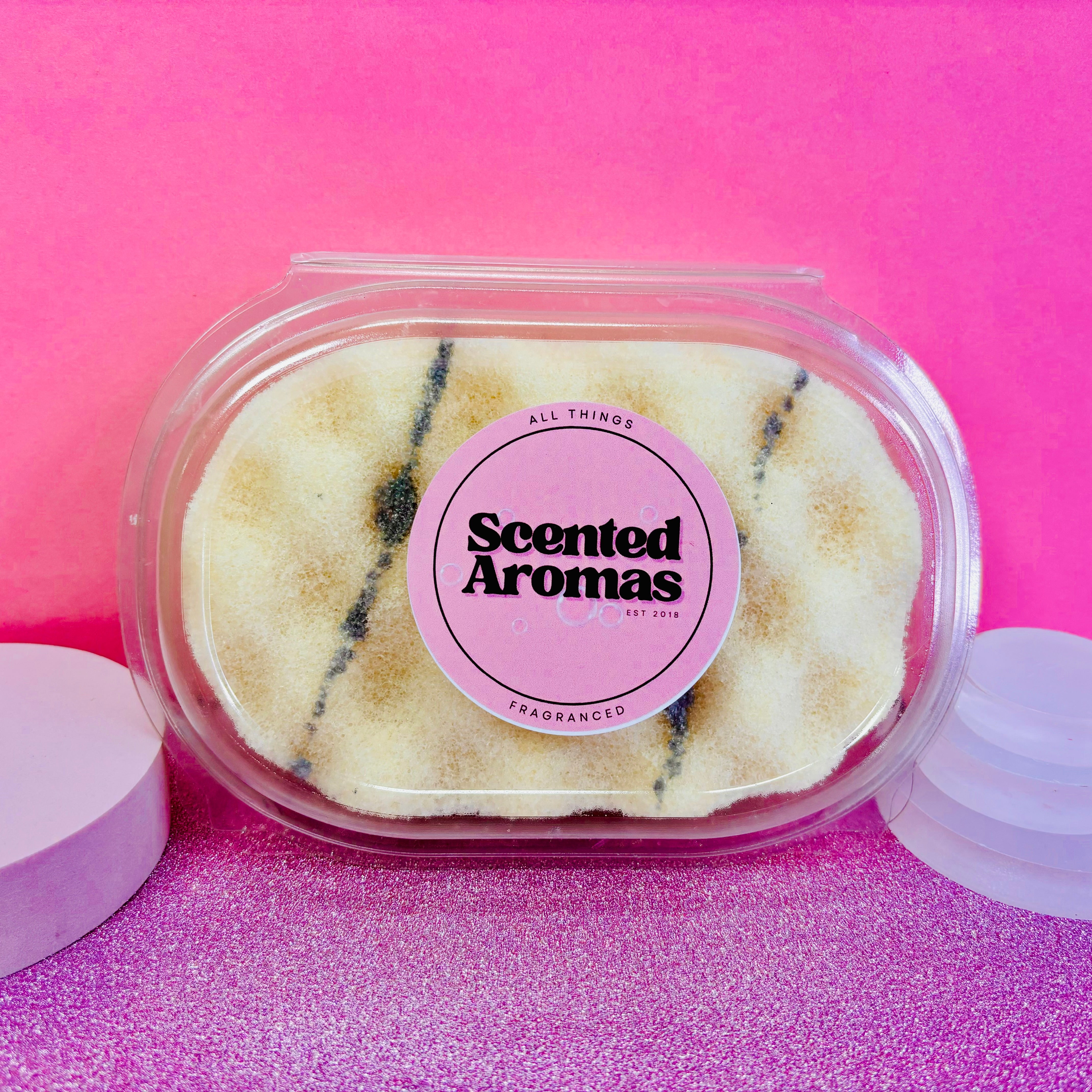 A plastic container labeled with "The Soap Gal x" holds a Monkey Farts Exfoliating Soap Sponge, featuring a white and brown striped design. It's set against a pink and glittery background, making it perfect for any skincare routine.