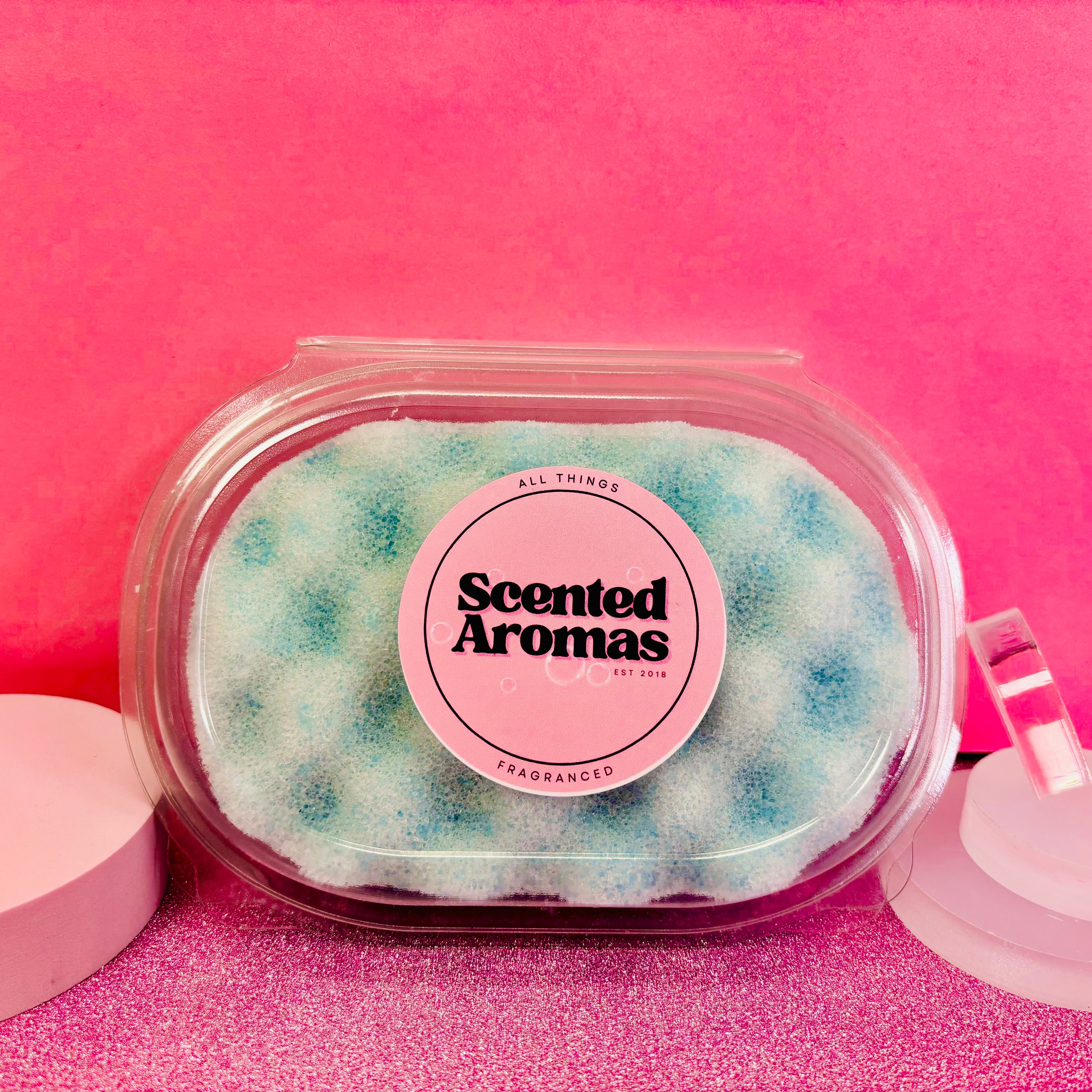 A container labeled "Savage Exfoliating Soap Sponge" by The Soap Gal x holds blue EVA foam beads, reminiscent of Sauvage notes, set against a pink background with two round white objects on either side.