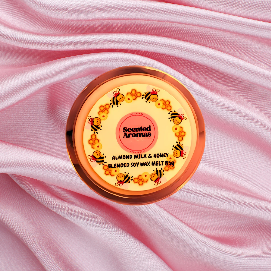 The Soap Gal x presents the Almond Milk and Honey Wax Melts (85g), packaged in a round container complete with a "Scented Aromas" label. Adorned with charming cartoon bees and flowers, this handmade wax melt sits gracefully on pink satin fabric, infusing any room with whimsy and warmth.