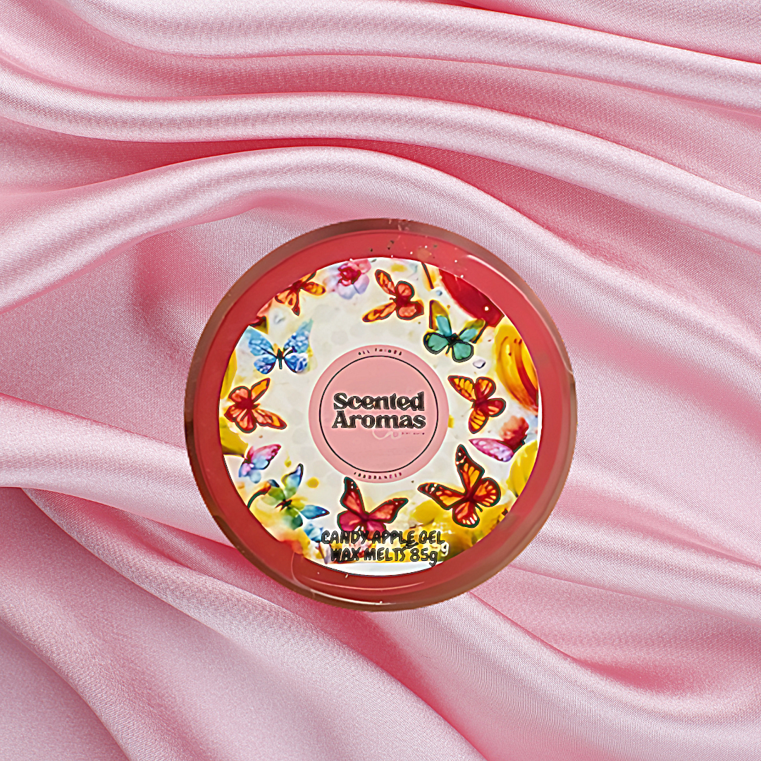 A round container labeled "The Soap Gal x" rests gracefully on pink satin fabric, its lid adorned with colorful butterfly designs. A delightful addition to your home accessories, it complements any décor with the enchanting fragrance of Frosted Candy Apple Gel Wax Melts 85g.