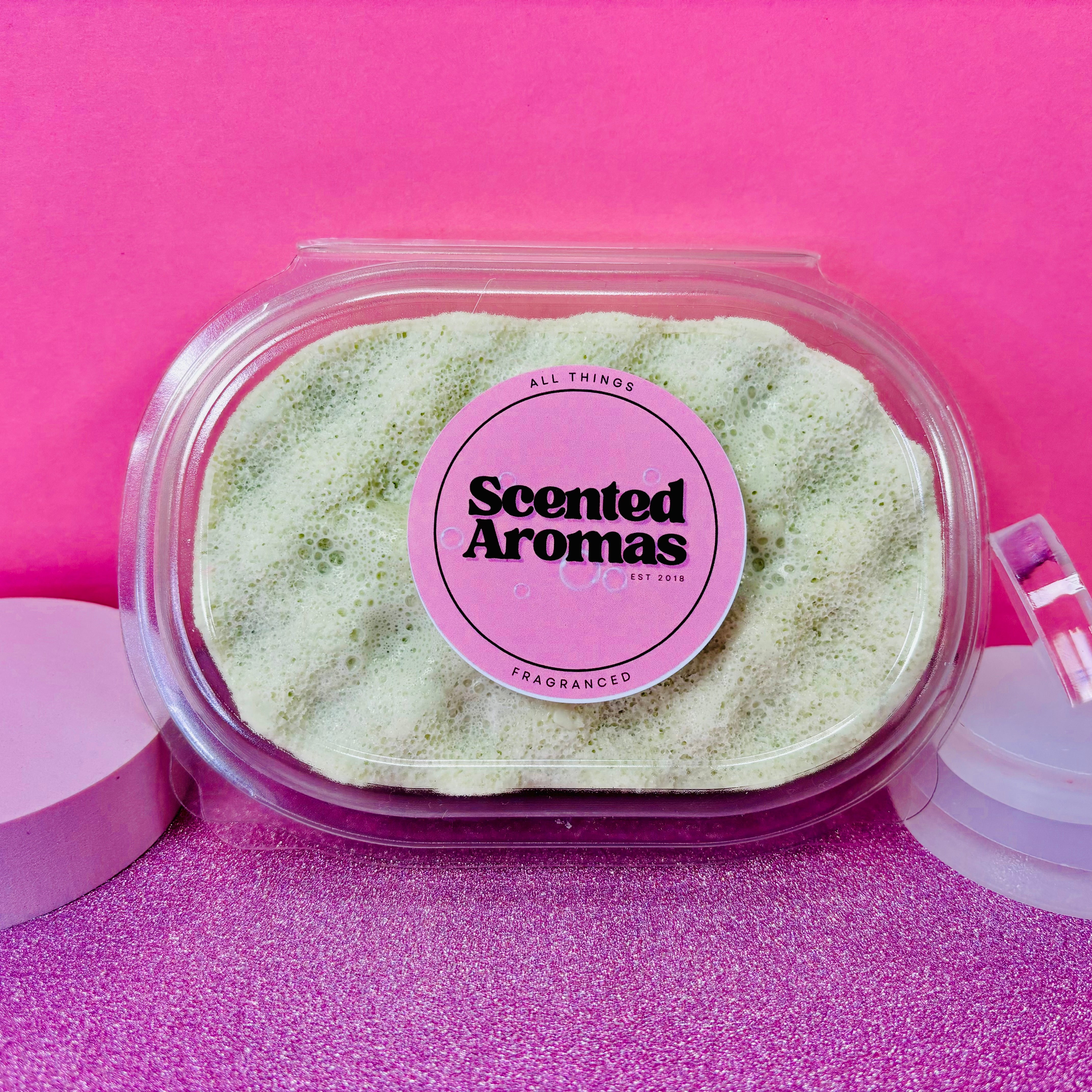 A Scented Aromas Alien Encounter Exfoliating Soap Sponge, featuring a pink label, is set against a vibrant pink background, offering an inviting scene of long-lasting fragrance and tactile exfoliating experience.