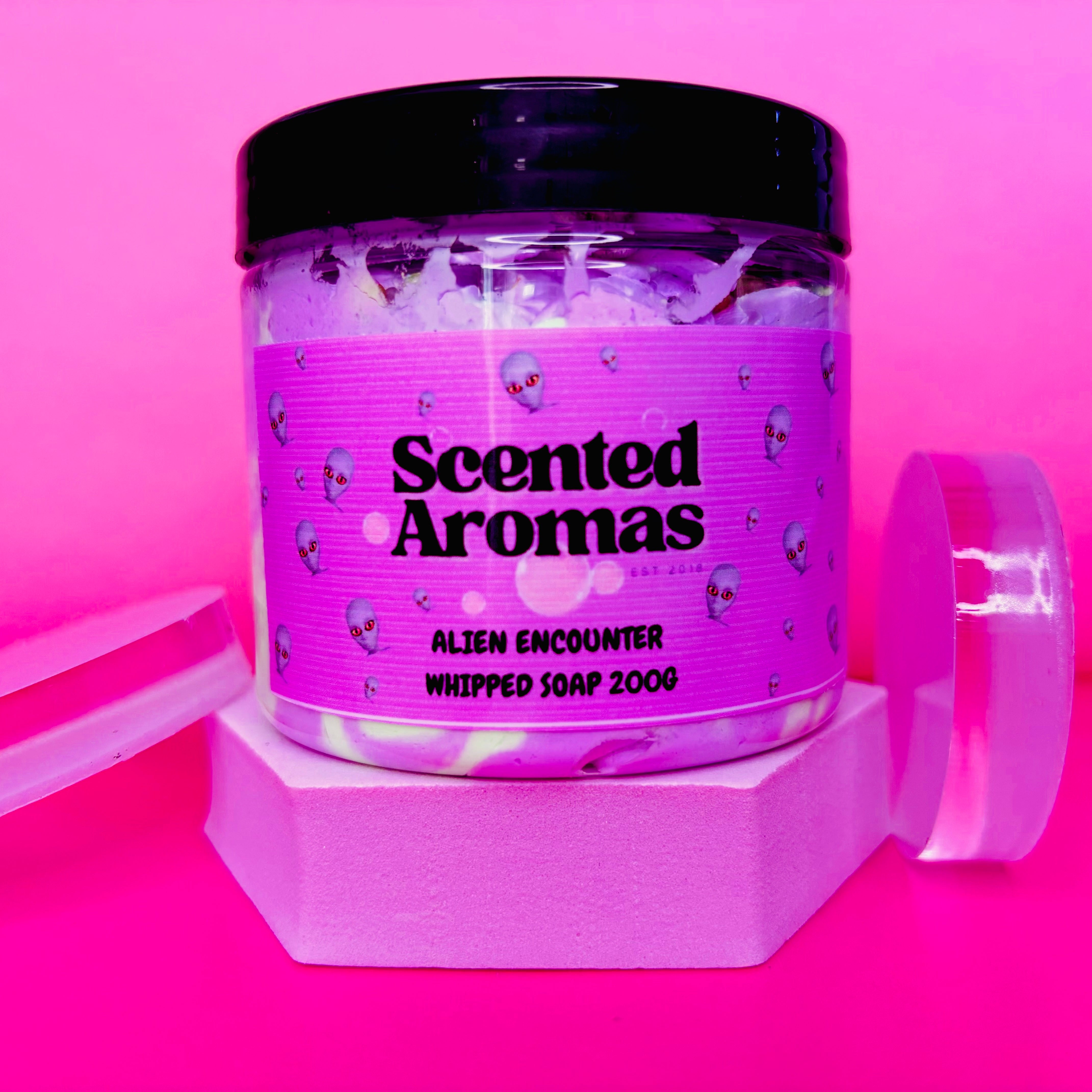 A jar of Scented Aromas' Alien Encounter Whipped Soap Shave Butter 200g, famous for its iconic scent, elegantly adorns a pink background with a hexagonal stand and lid beside it.