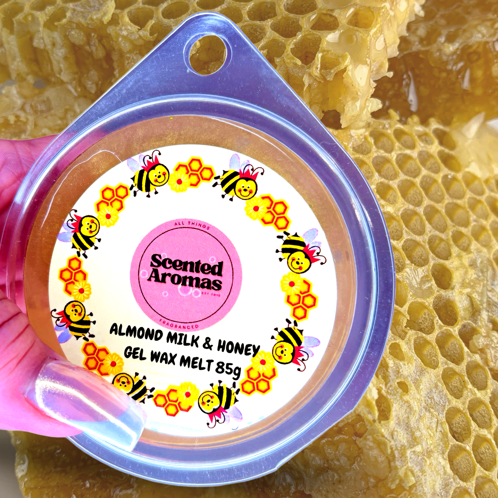 Close-up of a hand holding Scented Aromas' Almond Milk and Honey Gel Wax Melts 85g. The container showcases bee and honeycomb illustrations, set against a backdrop of natural honeycombs.