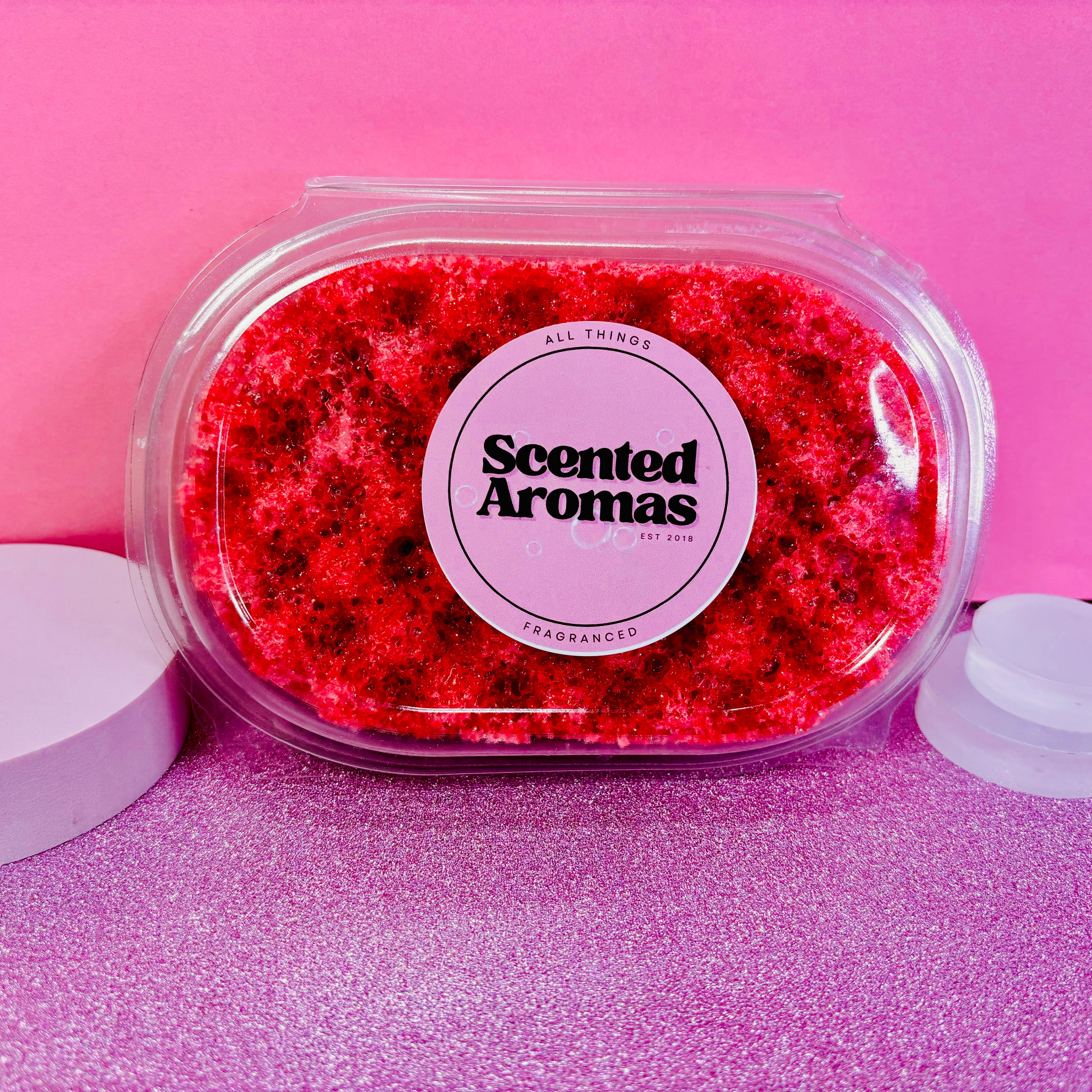 A Baccarat Rouge Exfoliating Soap Sponge from The Soap Gal x, featuring a red, scented soap in a clear oval container labeled "Scented Aromas," rests against a pink background, promising an indulgent skincare experience.