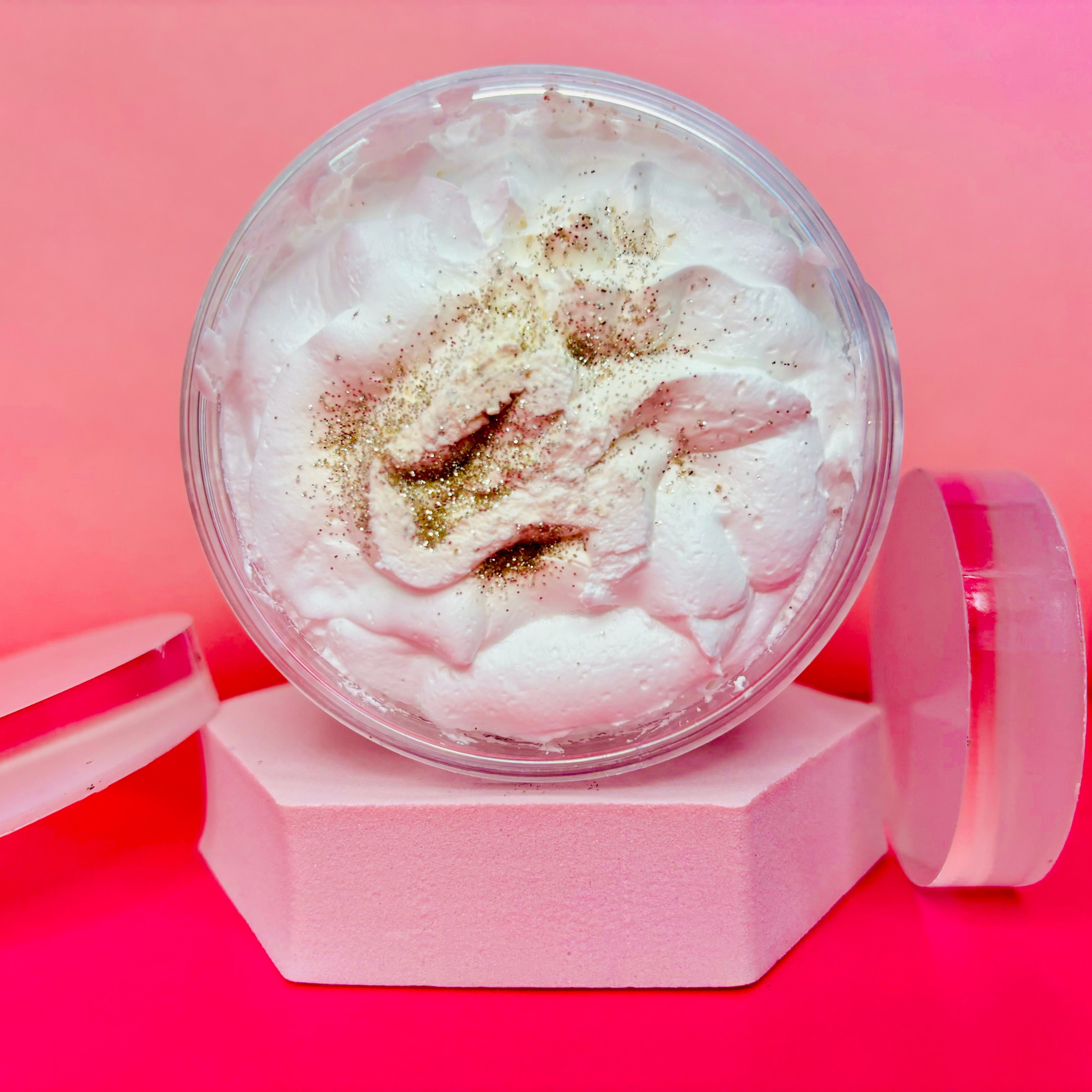 A round container of The Soap Gal x Almond Milk and Honey Whipped Soap Shave Butter 200g, adorned with white whipped cream and gold glitter, sits elegantly on a pink hexagonal platform. The vibrant pink background adds a touch of charm to this luxurious display, promising to tackle strawberry legs with ease.