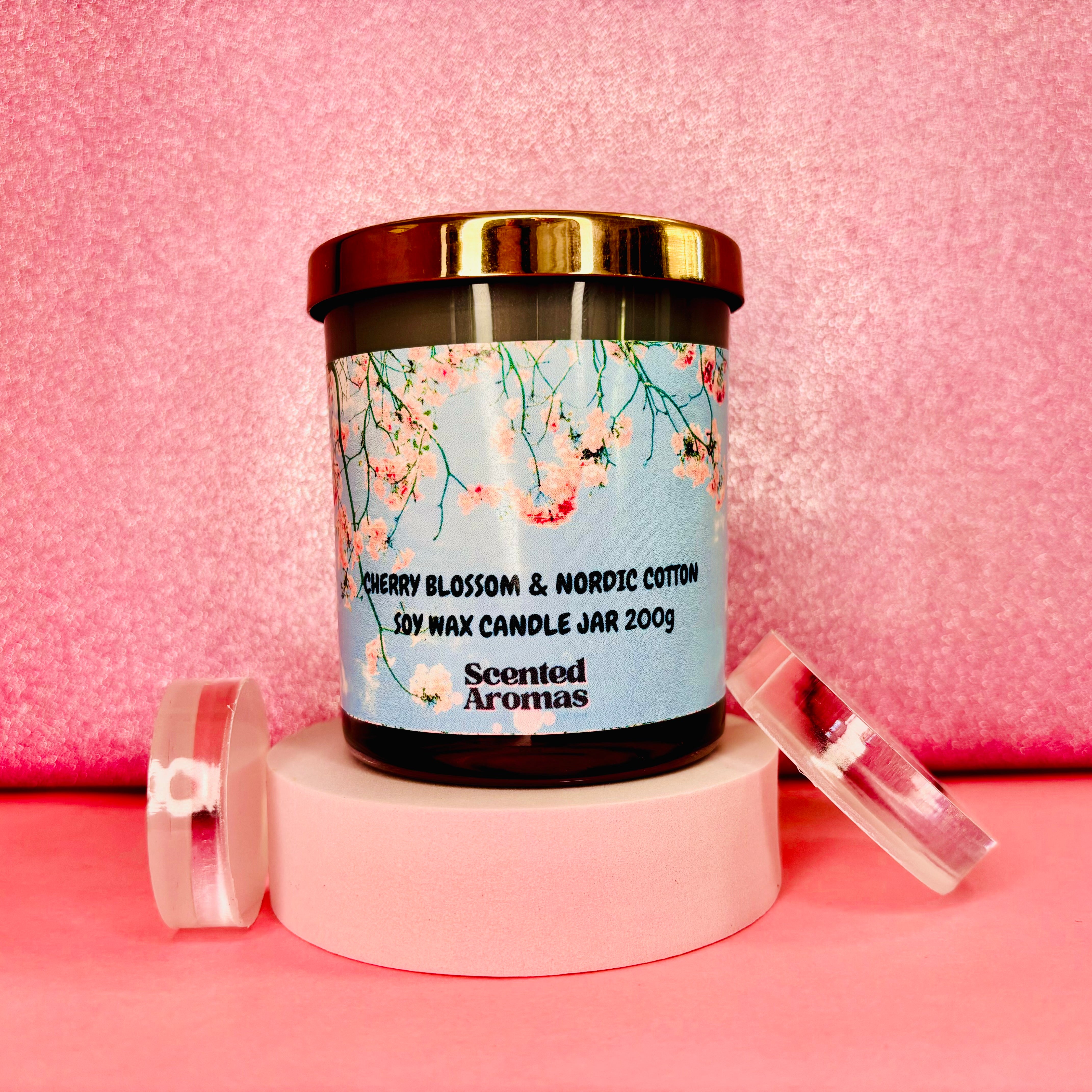 A jar candle labeled "The Soap Gal x Cherry Blossom and Nordic Cotton Candle 200g" sits on a pink background. This candle boasts floral artwork and a gold-colored lid, providing an elegant touch to any setting.