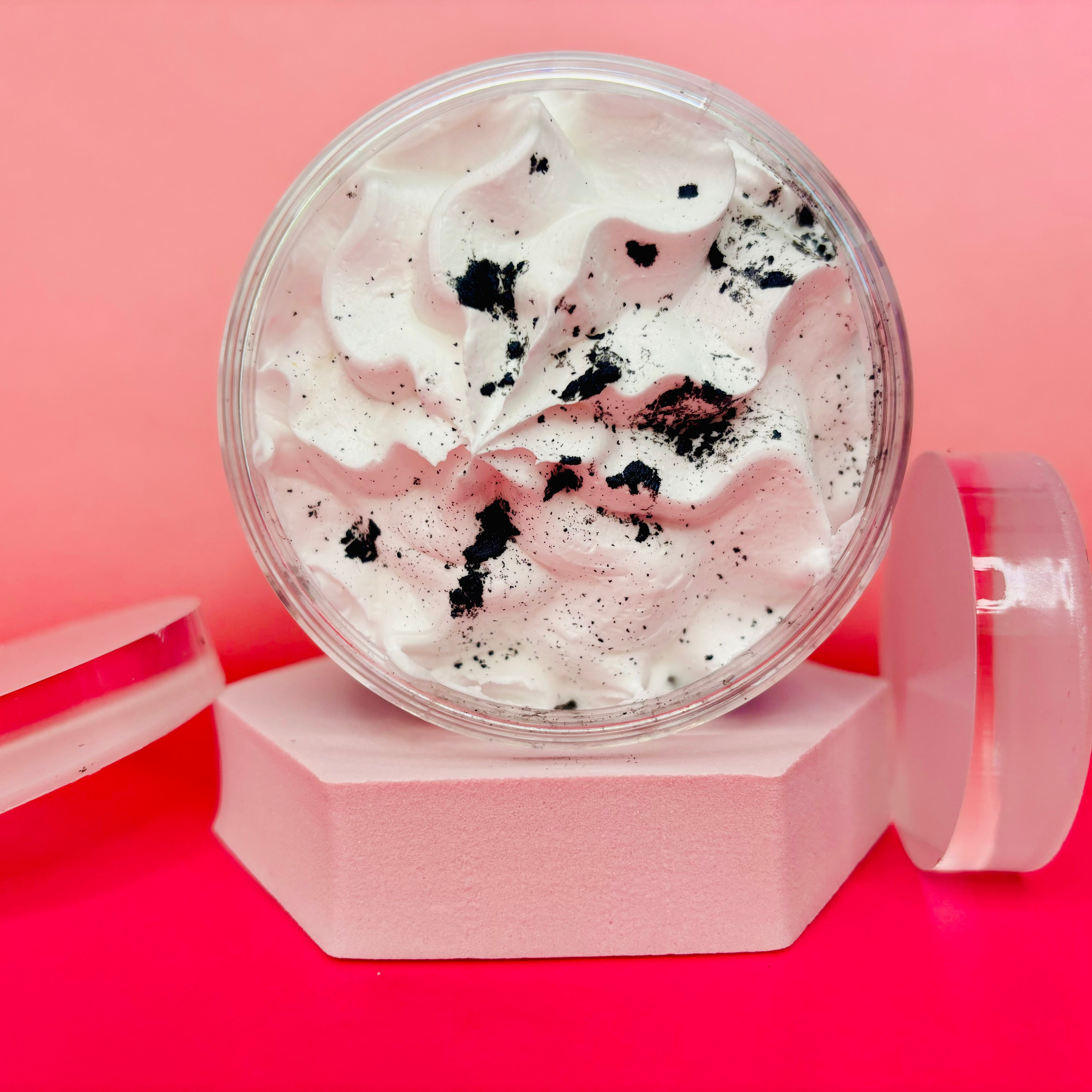 A container of Black Coconut Whipped Soap Shave Butter by The Soap Gal x, with its clear design showcasing the creamy texture and dark specks reminiscent of luxurious shave butter, is elegantly placed on a pink hexagonal platform set against a dual-toned pink background. The matching lid leans effortlessly against the container, reflecting the sophistication of high-end skincare routines.