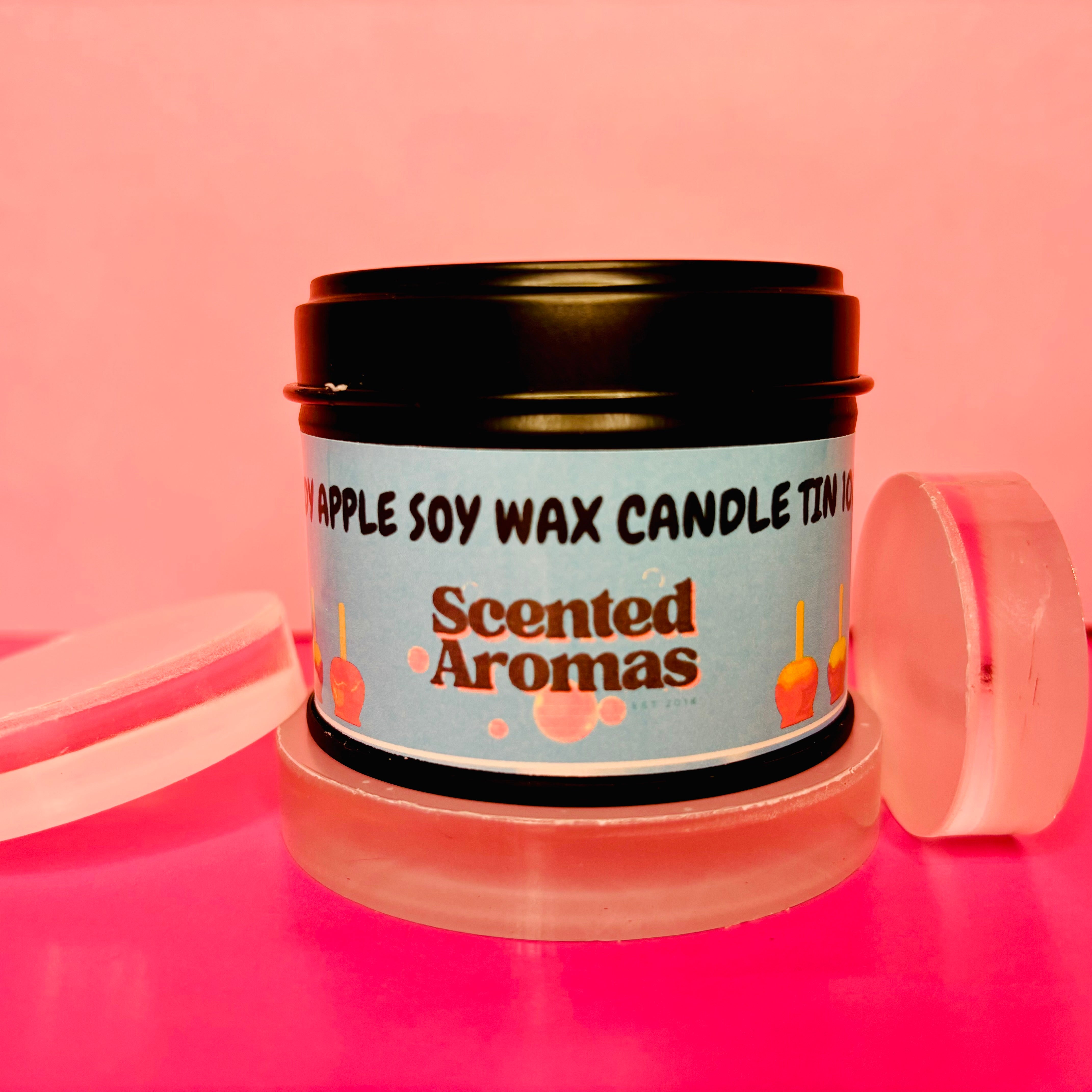 An Apple Candy Candle Tin by The Soap Gal x rests on a pink background, with the travel-friendly lid set aside. Made from soy vegan-friendly wax, this delightful 100ml candle adds elegance to any setting.