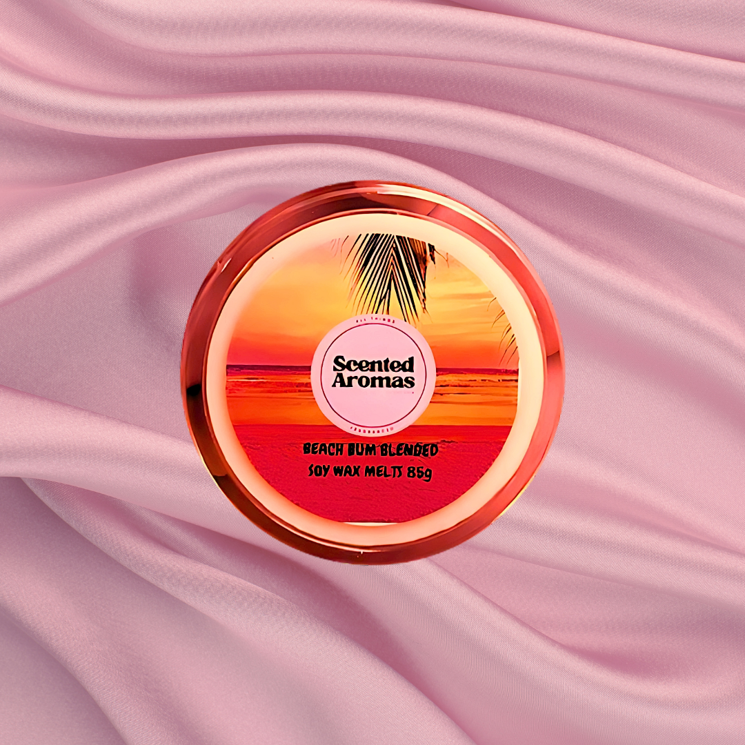 A round container labeled "The Soap Gals Brazilian Bum Cream Wax Melt 85g" rests on a pink, silky fabric background, reminiscent of the luxurious feel of beach-themed products.