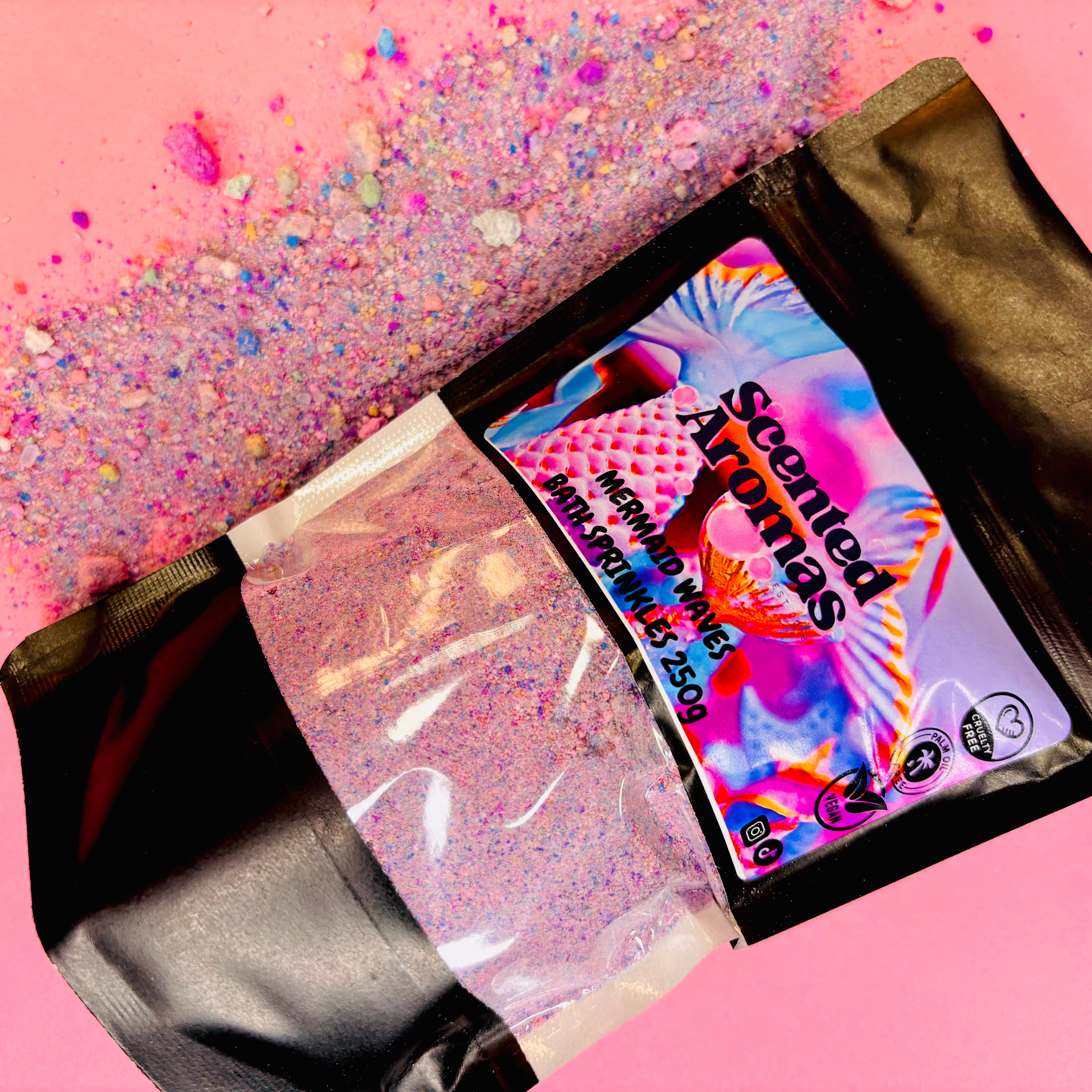 A colorful package labeled "The Soap Gal x" sits on a pink surface, with vibrant Mermaid Waves Bath Fizzing Salts granules scattered beside it, releasing a hint of Tooti Fruity fragrance.