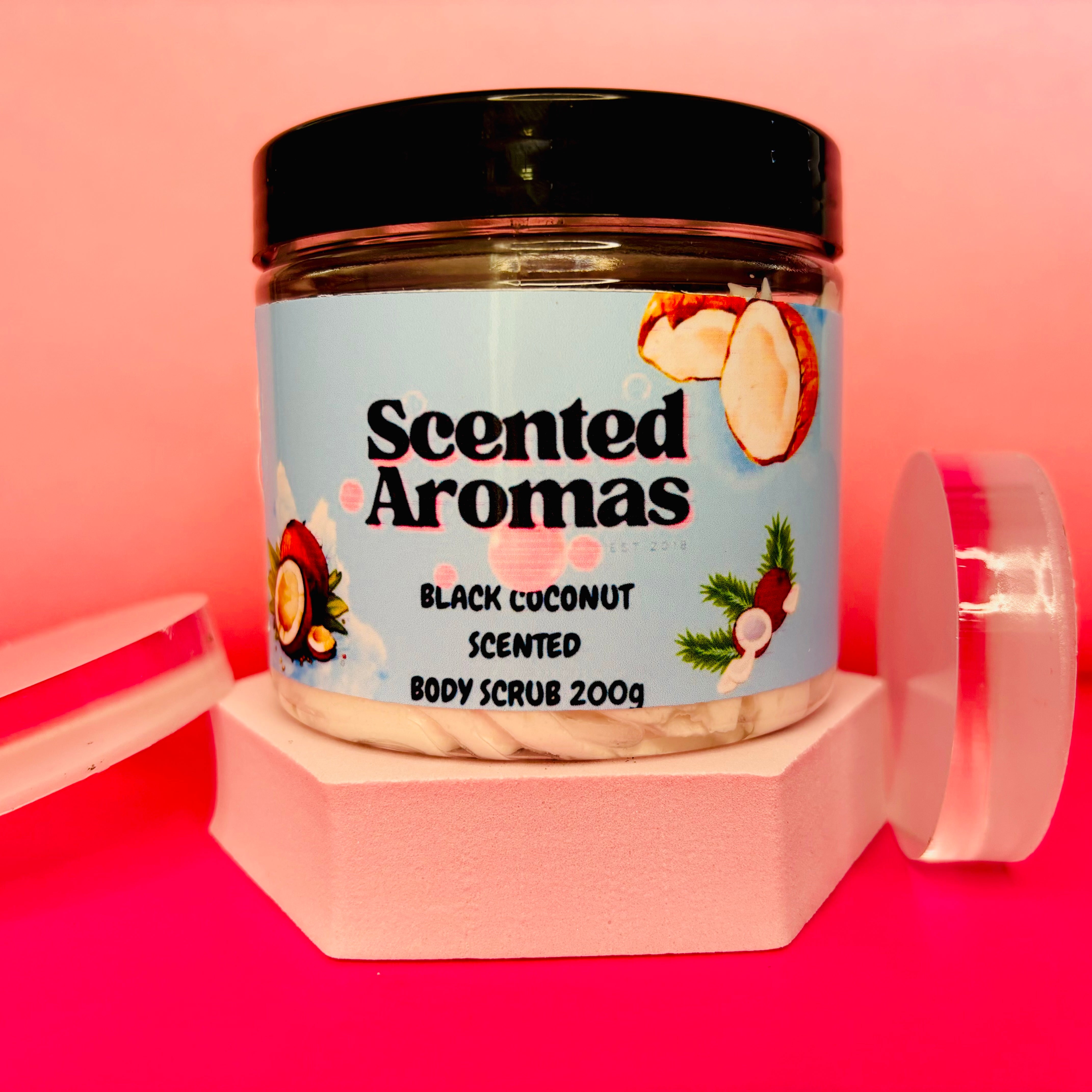 A 200g jar of Black Coconut Sugar Body Scrub by The Soap Gal x provides a spa-like experience on a pink platform, set against a pink background.