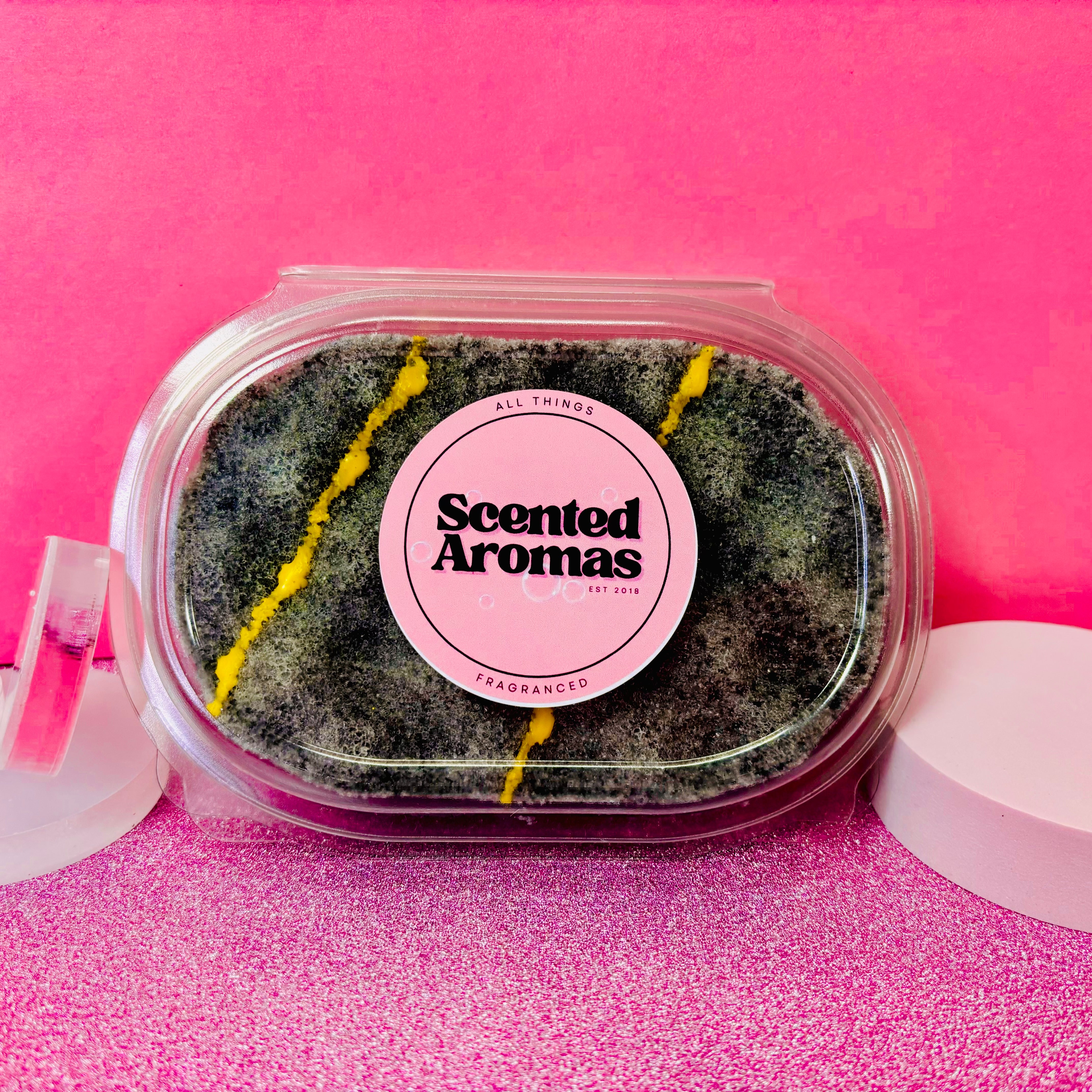 An Invictus Exfoliating Soap Sponge by The Soap Gal x, with a masculine fragrance and black and yellow contents, rests on a glittery pink surface against a pink background.