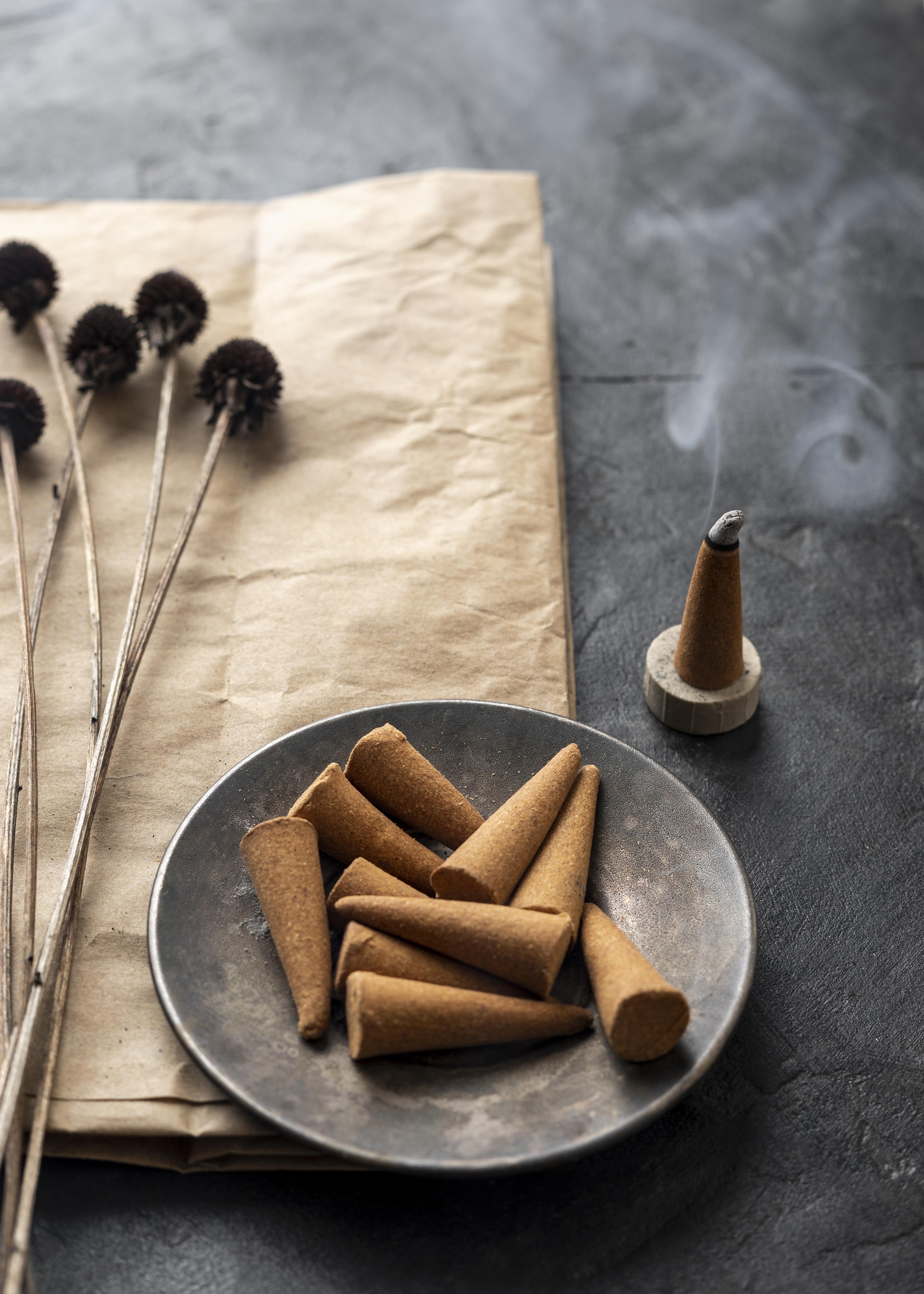 A collection of Nag Champa Incense Cones from The Soap Gal x sits on a plate, with one gracefully smoking in an elegant holder. Artistic arrangements of dry seed pods decorate the nearby brown paper.