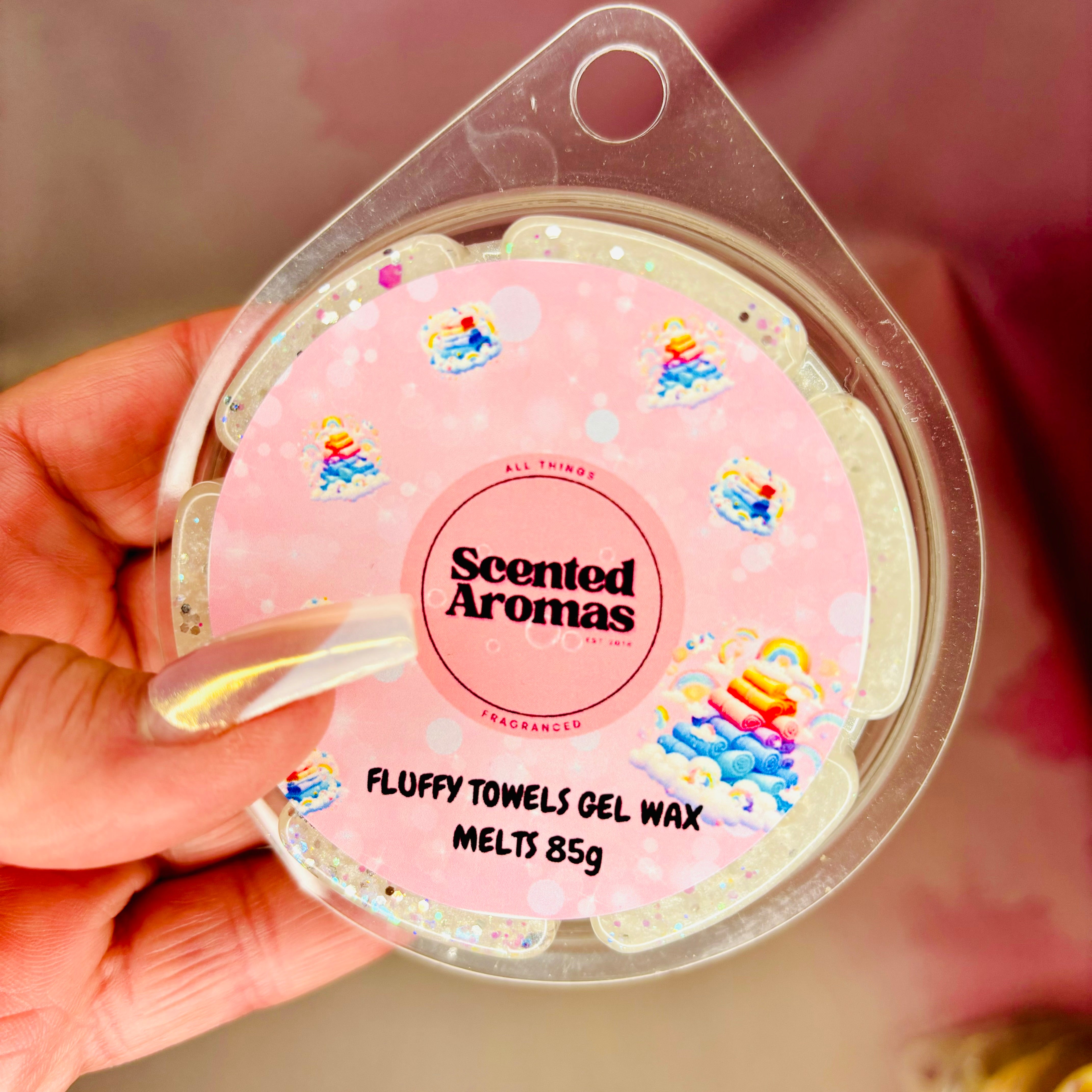 A hand displays a container of The Soap Gal x Fluffy Towels Gel Wax Melts, 85g, against a pink background with vibrant illustrations. Enjoy the unique fragrance blends that set these vegan wax melts apart.