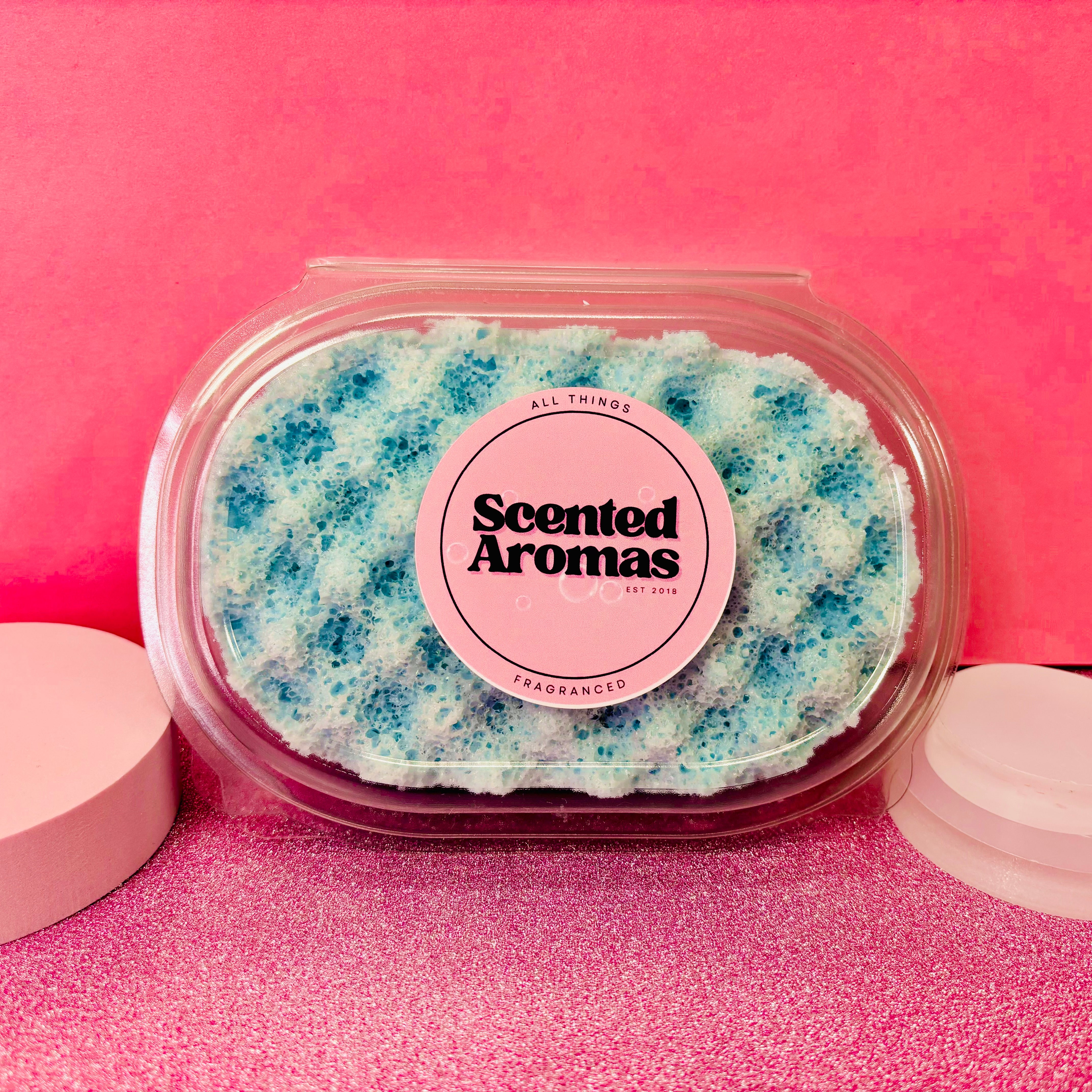 A Sea Mint Minerals Exfoliating Soap Sponge by The Soap Gal x, featuring a blue color with white accents, packaged in plastic and labeled "Scented Aromas," set against a pink background, perfect for your skincare routine.