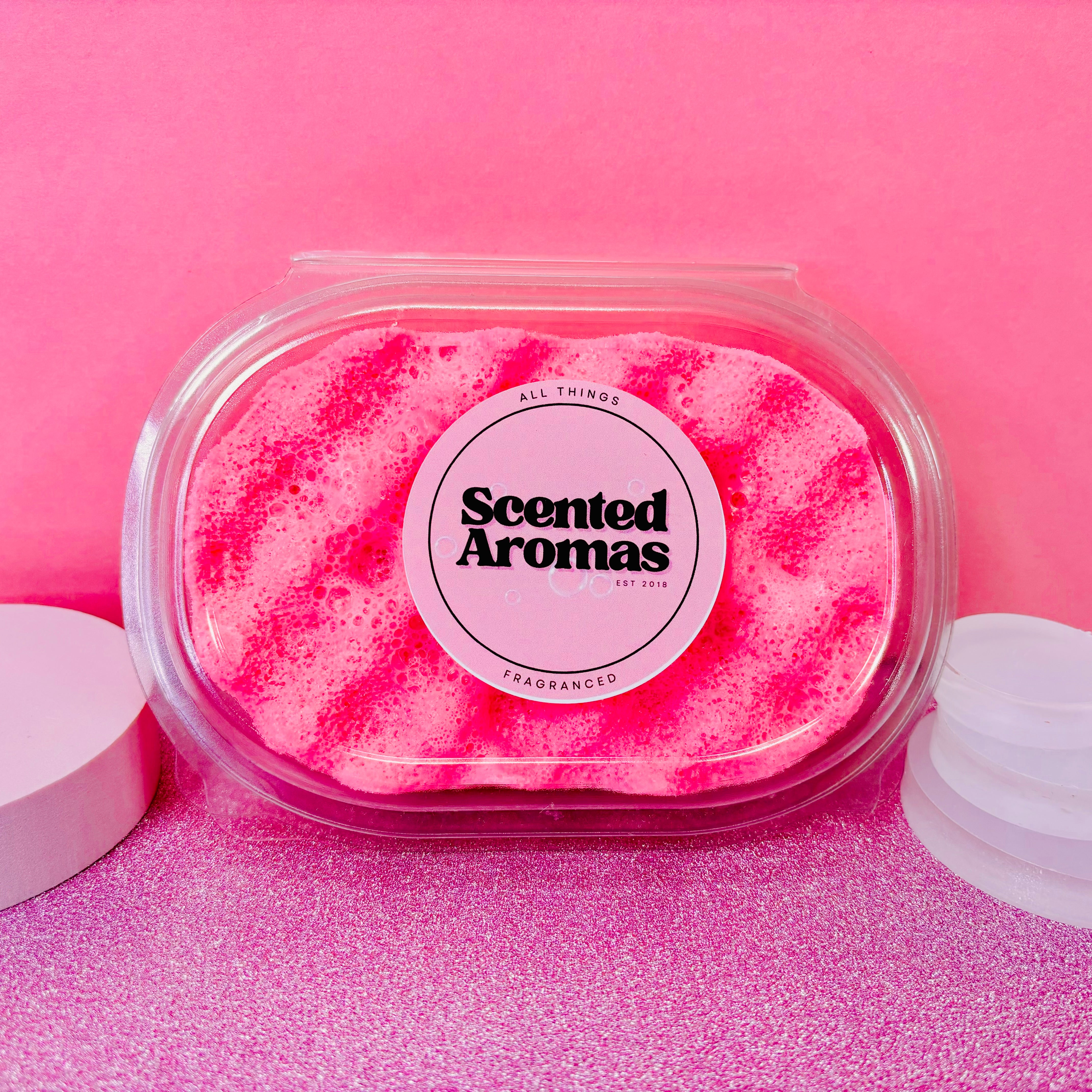 The "Fantasy Exfoliating Soap Sponge" by The Soap Gal x, featuring a pink hue and captivating fragrance, is showcased in a clear plastic container against a pink backdrop. This handmade creation from Manchester promises an enchanting journey through delightful aromas.