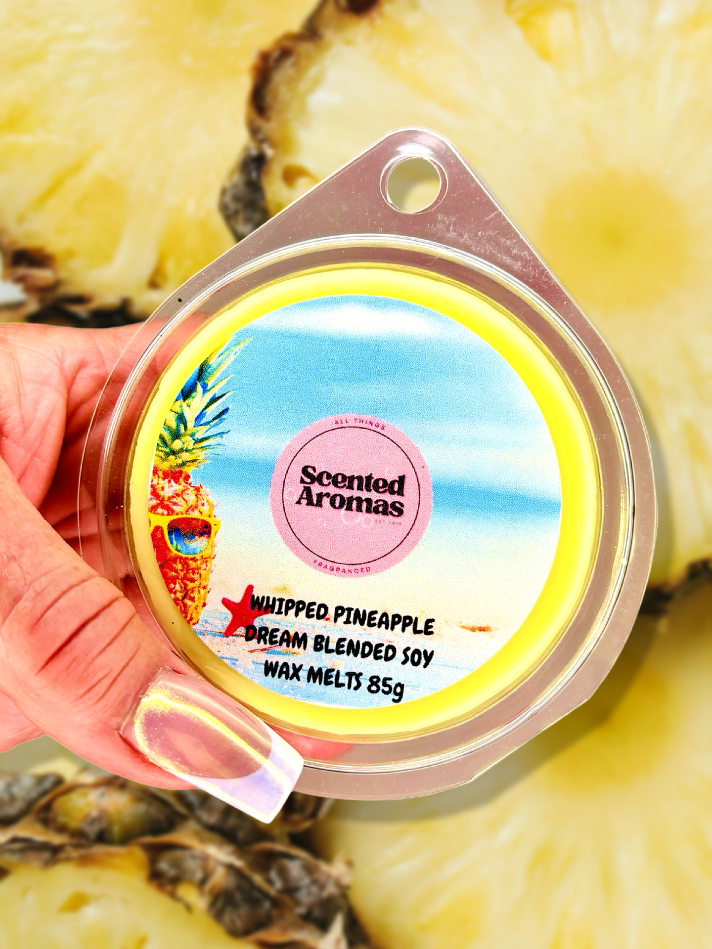 A hand holds a "The Soap Gal x" Pineapple Whip Wax Melts 85g container, spreading a fruity ambiance. The background features sliced pineapples.