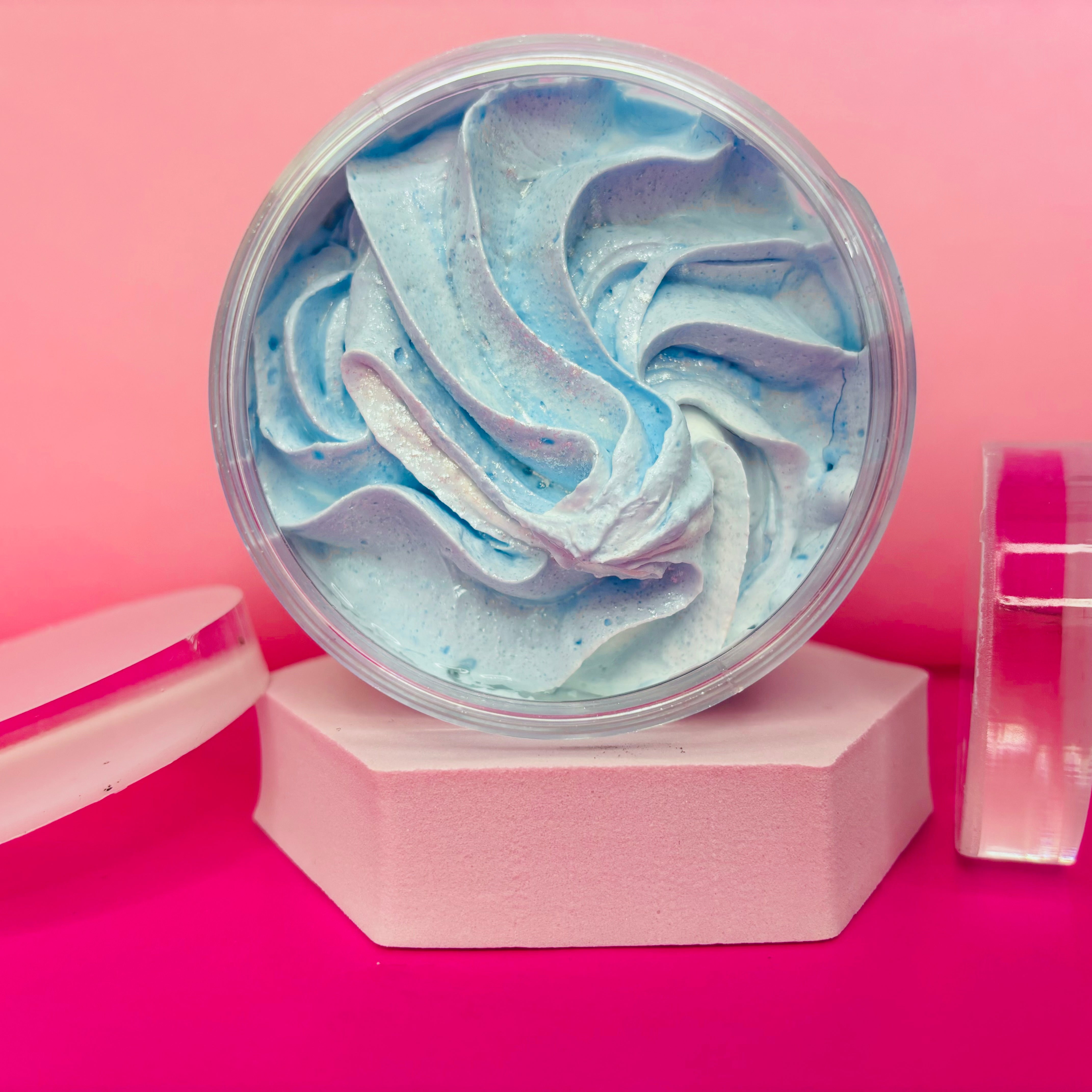 The Soap Gal x Snow Pixie Sugar Body Scrub 200g is presented in a round container filled with swirled blue and white cream, set against a pink and red backdrop on a hexagonal platform, accompanied by a clear lid. This vegan sugar scrub offers an indulgent exfoliating experience.