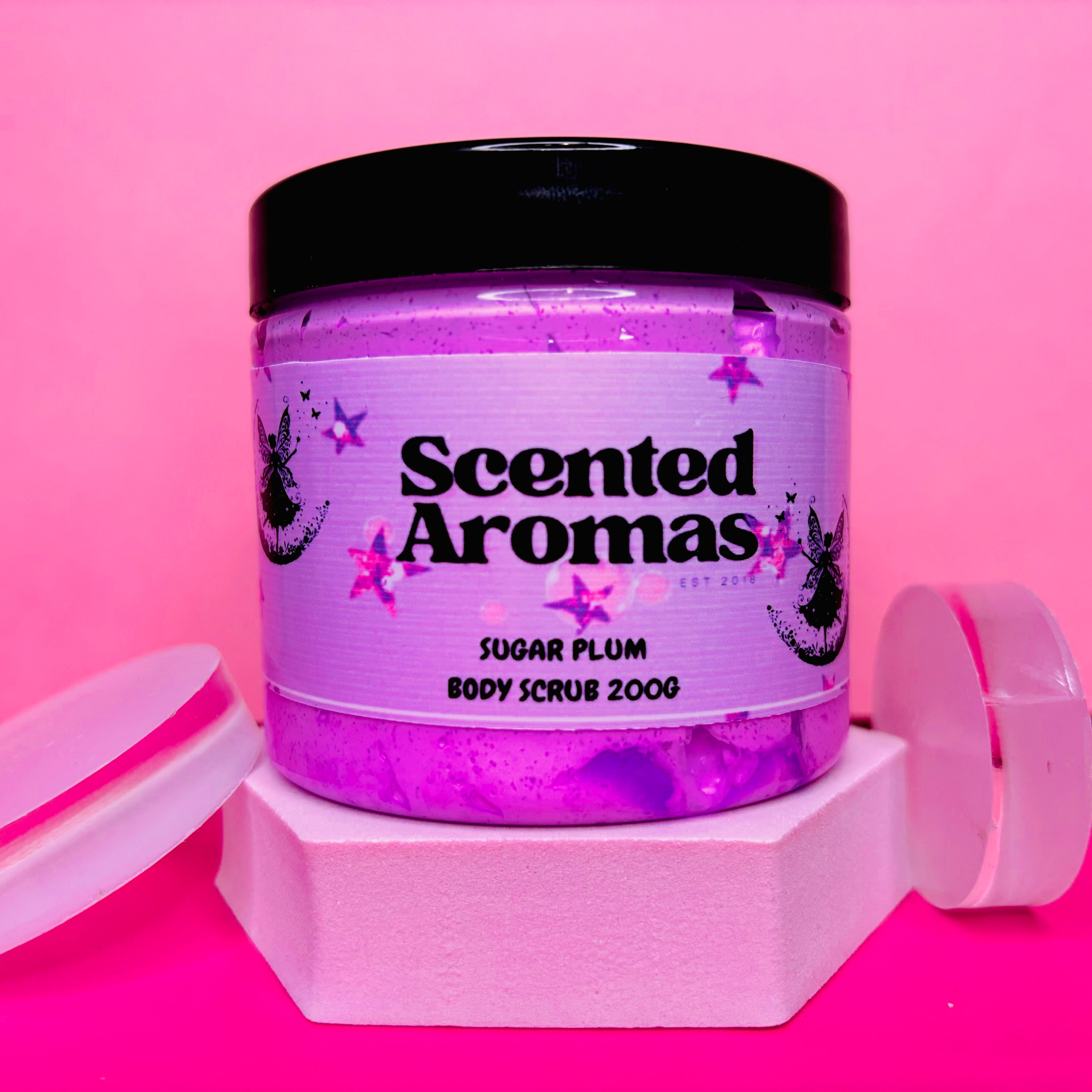 A jar of The Soap Gal x Frosted Sugar Plum Sugar Body Scrub, featuring a black lid, rests on a pink background, promising smooth skin.