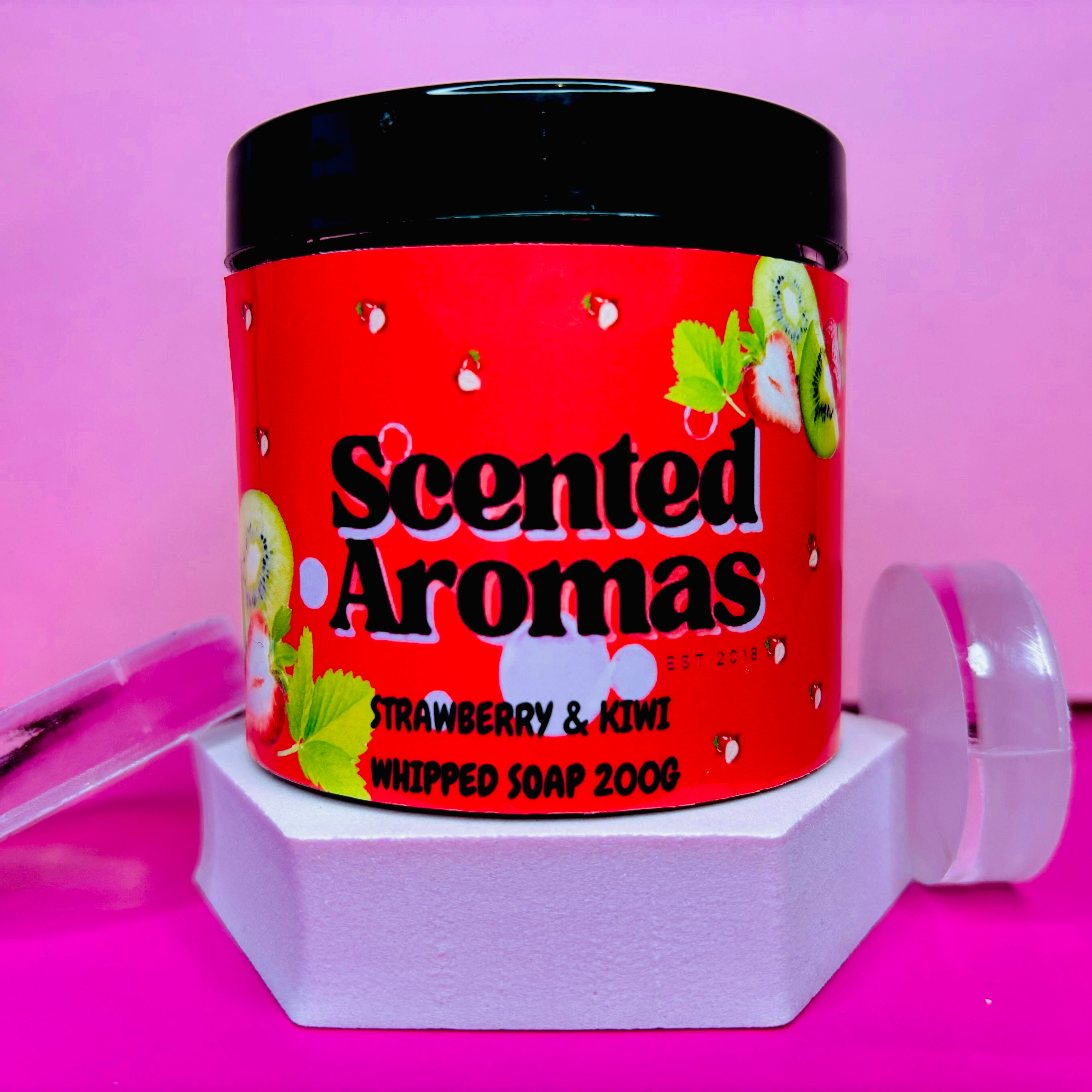 A jar of "The Soap Gal x" Strawberry and Kiwi Whipped Soap Shave Butter, 200g, elegantly rests on a white hexagonal stand against a pink background, promising a luxurious shaving experience.