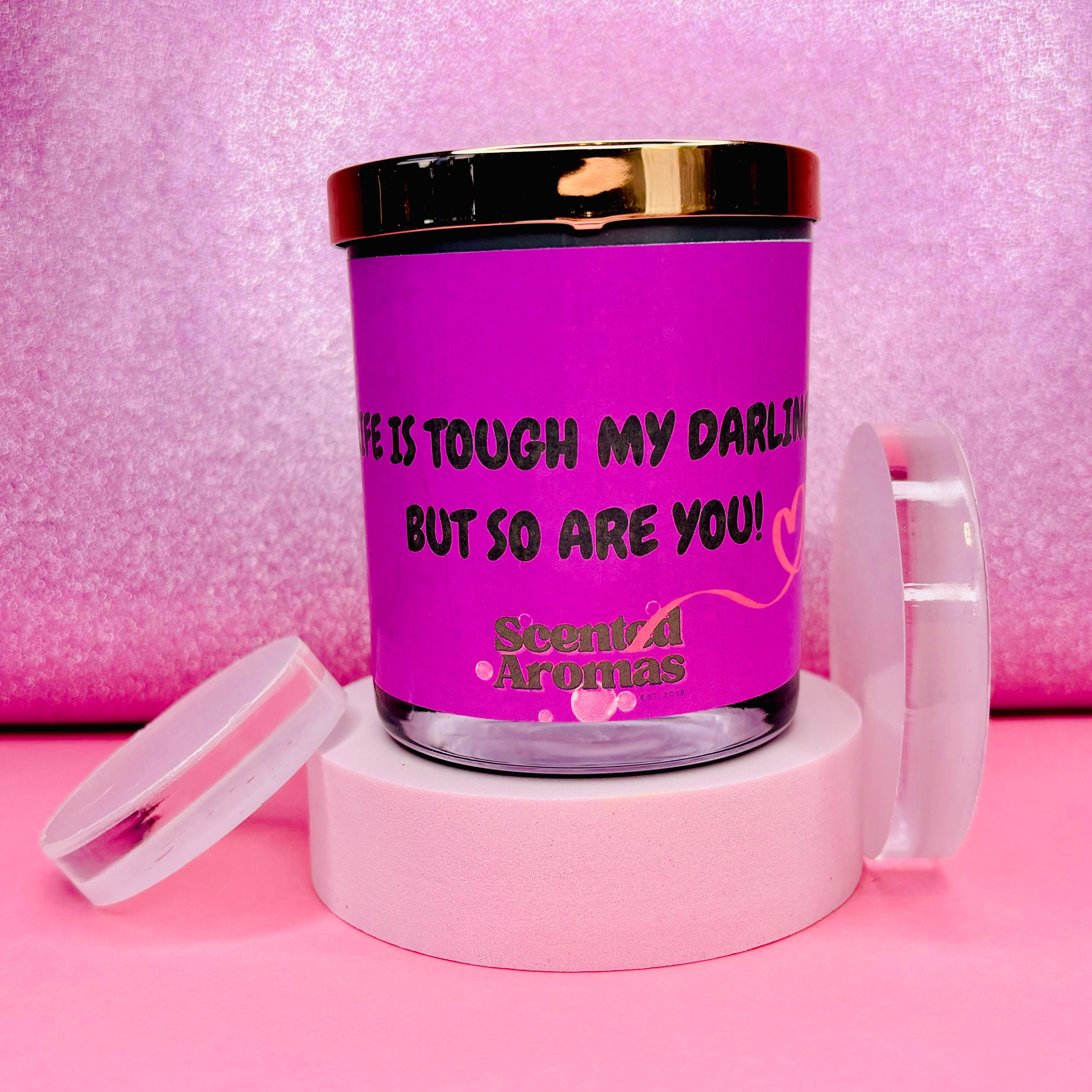 The Life is Tough Positive Affirmation Quote Candle 200g by The Soap Gal x features a pink design and sits elegantly on a white stand with its lid placed nearby. Crafted from 100% pure soy wax, it bears the uplifting message, "Life is tough my darling, but so are you!" providing a charming reminder of strength and resilience.