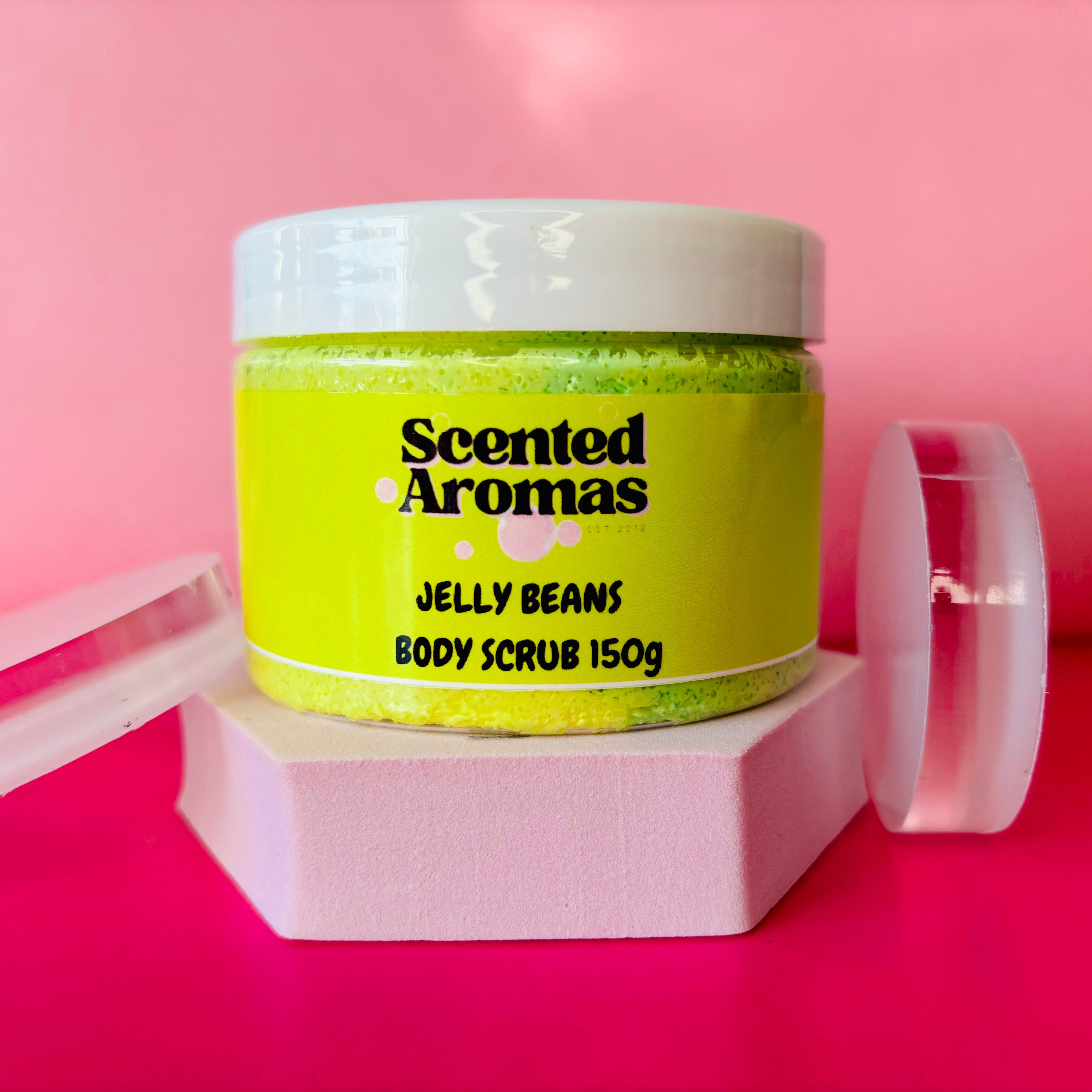 A jar of The Soap Gal x Jelly Beans Body Scrub 150g rests on a white hexagonal platform against a pink background, showcasing its yellow label. This exfoliating product offers a promise of smoother skin with every application.