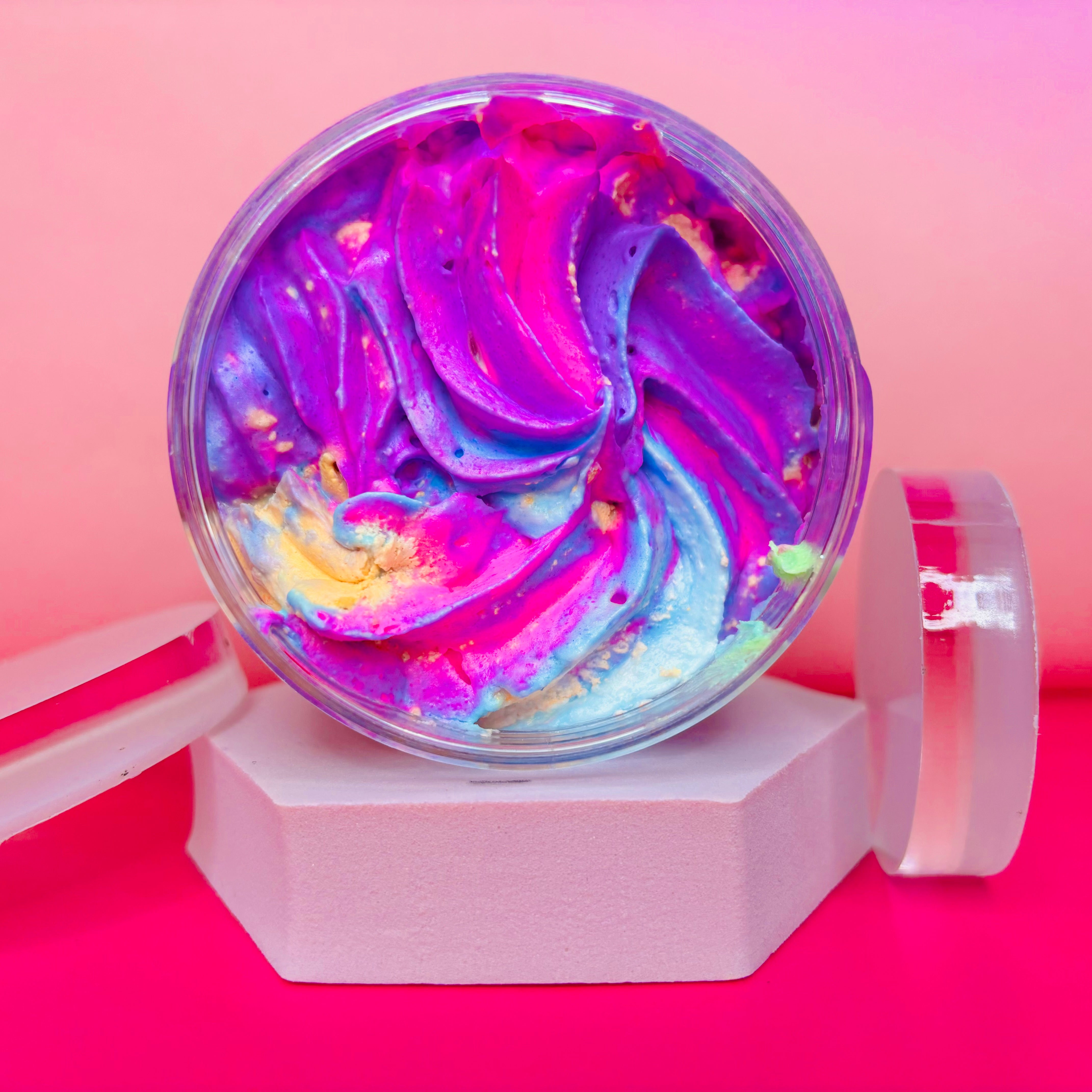 A jar of the vibrant Unicorn Fluff Whipped Soap Shave Butter 200g by The Soap Gal x, showcasing a mesmerizing blend of purple, pink, blue, and yellow swirls, rests on a white hexagonal stand against a gradient pink background. Its mix of colors and Tropical Colada scent offer a whimsical treat for your skin.