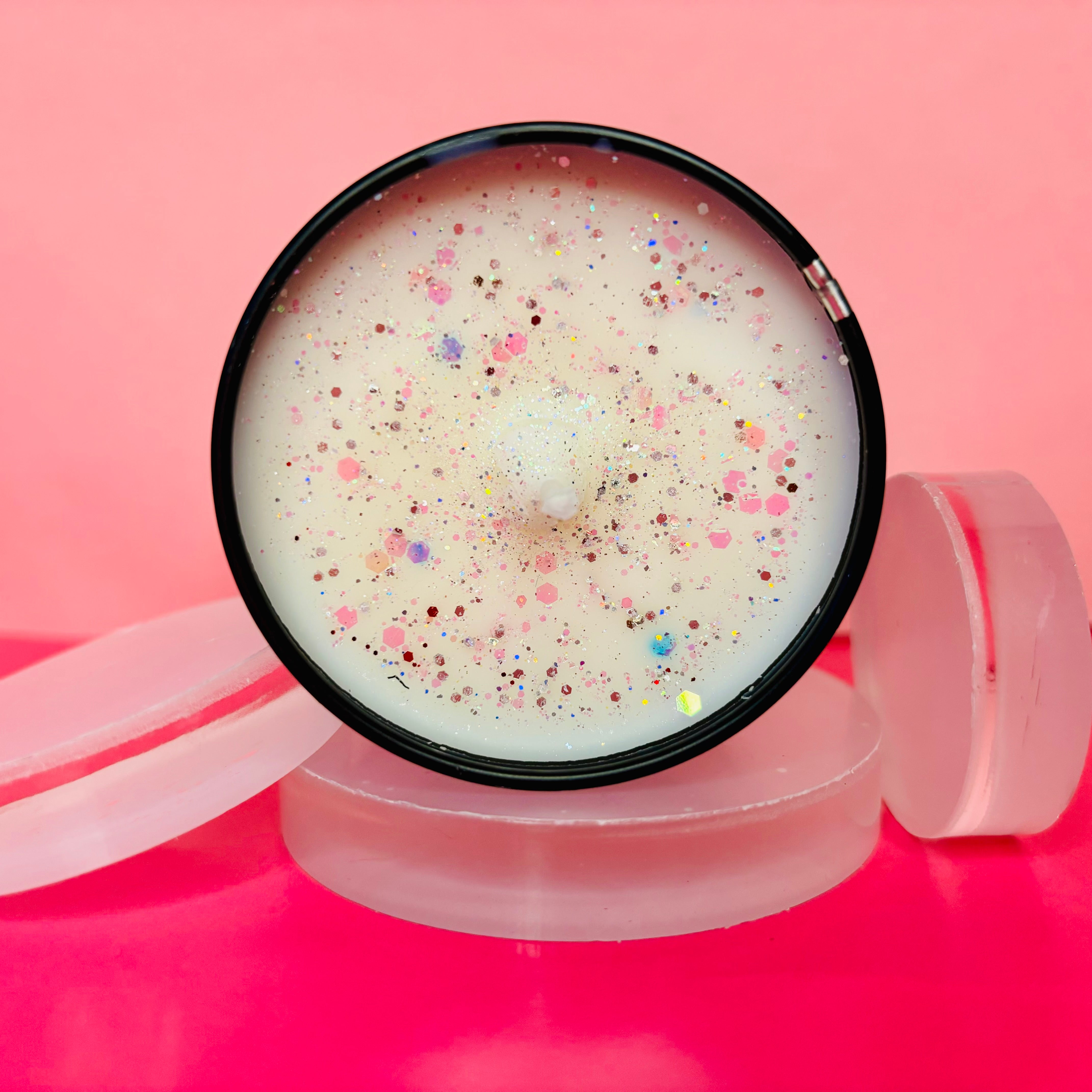 The Snow Pixie Candle Tin - 100ml from The Soap Gal x gleams with colorful glitter on its round surface. An open lid reveals two frosted discs inside, set against a pink background, capturing the enchanting essence of a vegan-friendly home fragrance.