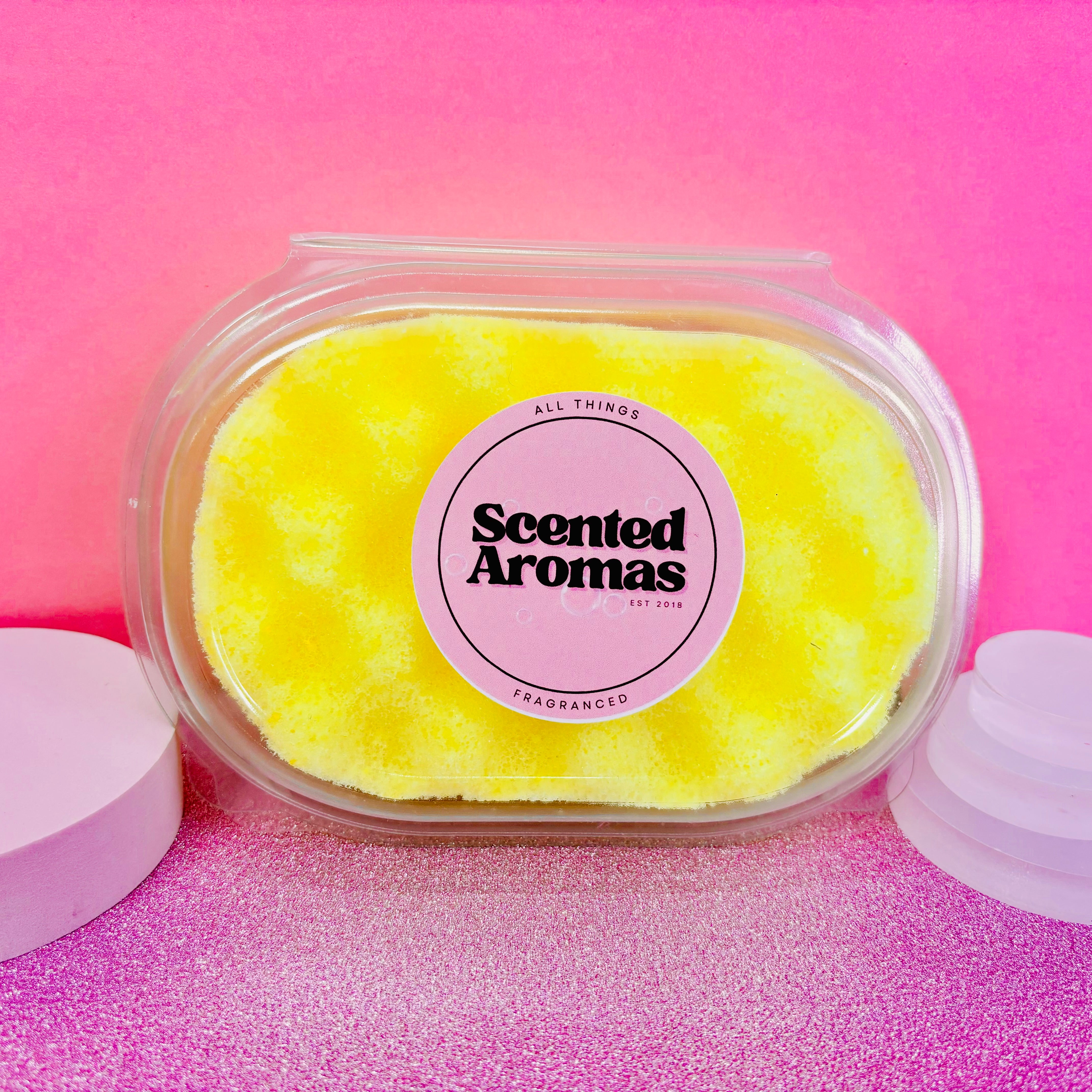 The yellow scented soap, called "Scented Aromas," is packaged in a plastic container set against a pink background. It is an excellent addition to your skincare routine and is crafted to complement the Coco Cabana Exfoliating Soap Sponge by The Soap Gal x.