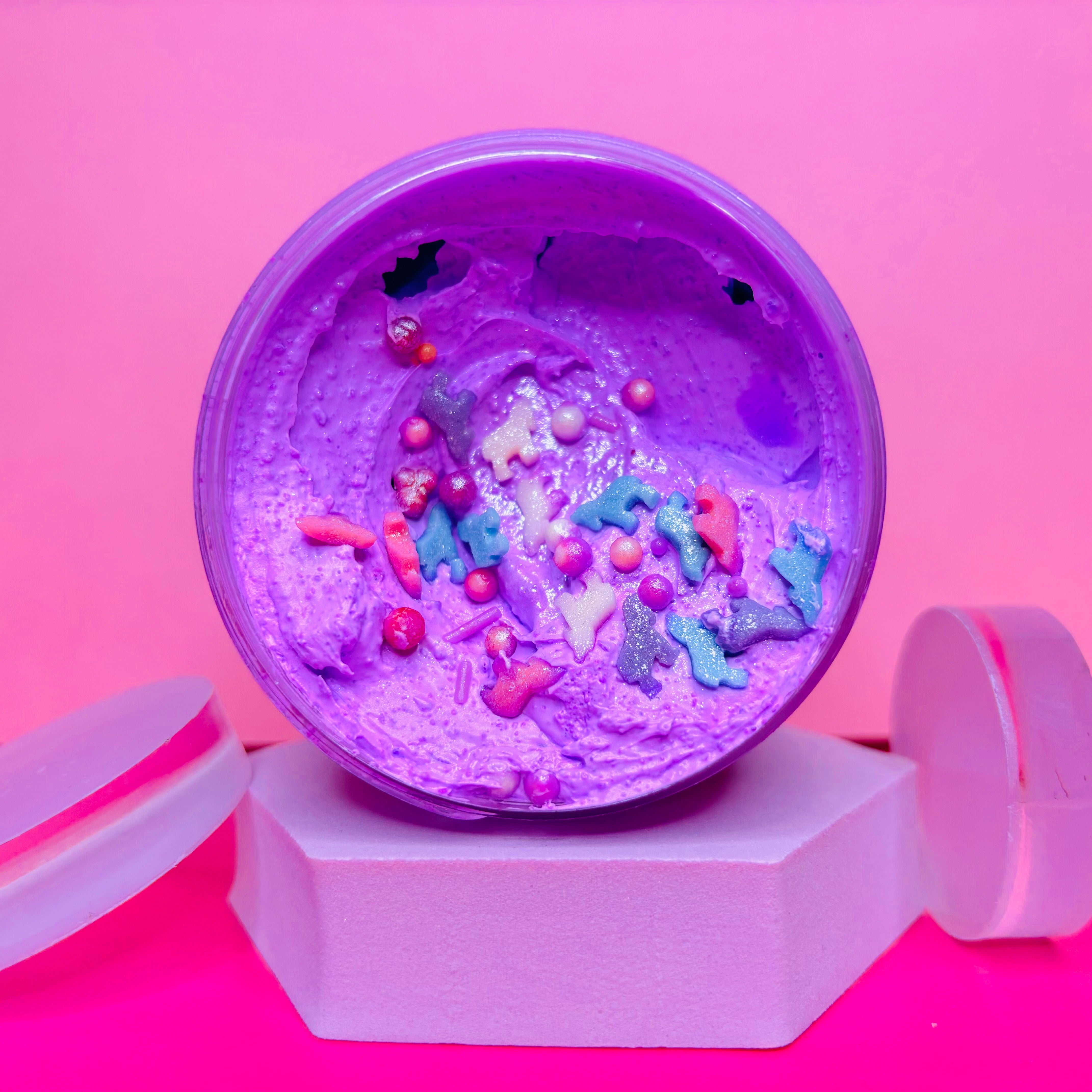 In a visually appealing setup, the Frosted Sugar Plum Sugar Body Scrub 200g by The Soap Gal x is showcased in a round container, adorned with colorful star and pearl decorations, resting on a white hexagonal stand against a pink background—perfect for achieving smoother skin.