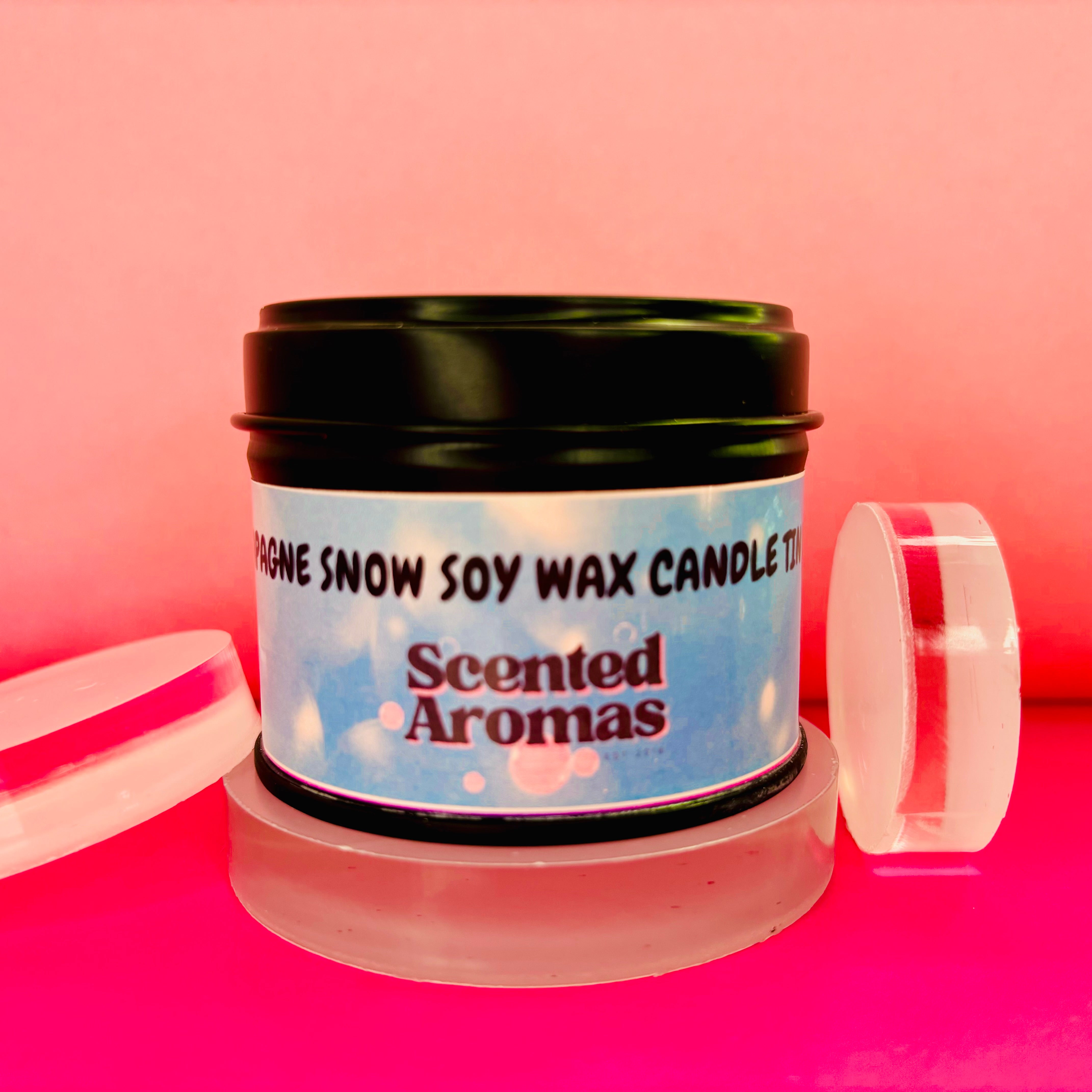 A tin of "Snow Champagne Candle - 100ml" by The Soap Gal x, infused with a delicate floral fragrance, sits elegantly on a red and pink background with two clear circular objects beside it.