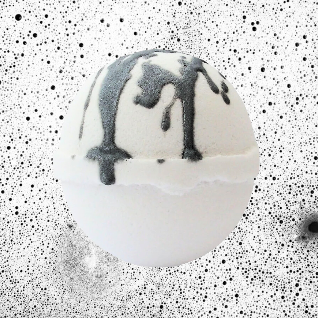 A spherical white Flowerbomb Bath Bomb from The Soap Gals, adorned with black drips, floats against a speckled, black-and-white dotted background, offering a luxurious vegan treat infused with the enchanting scent of Jasmine Sambac.