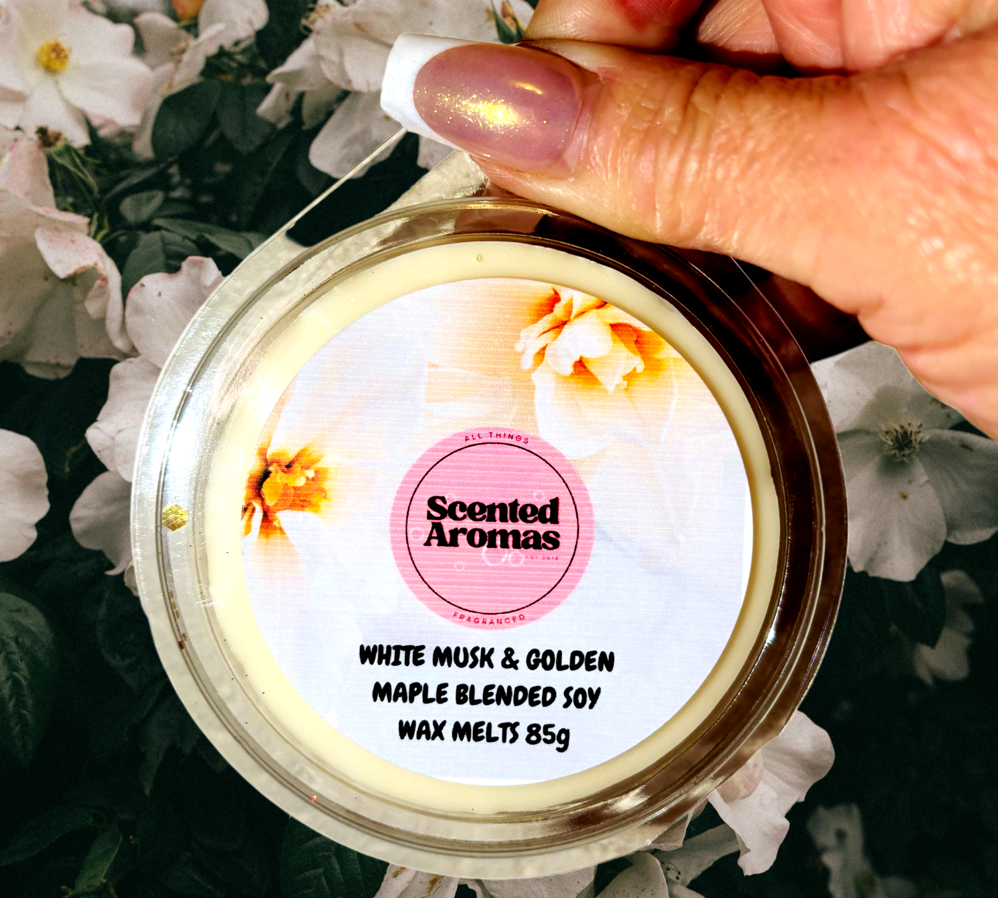 A hand holds an 85g container of Hinch's White Musk and Golden Maple Wax Melts by The Soap Gal x, with delicate white flowers providing a soft backdrop.