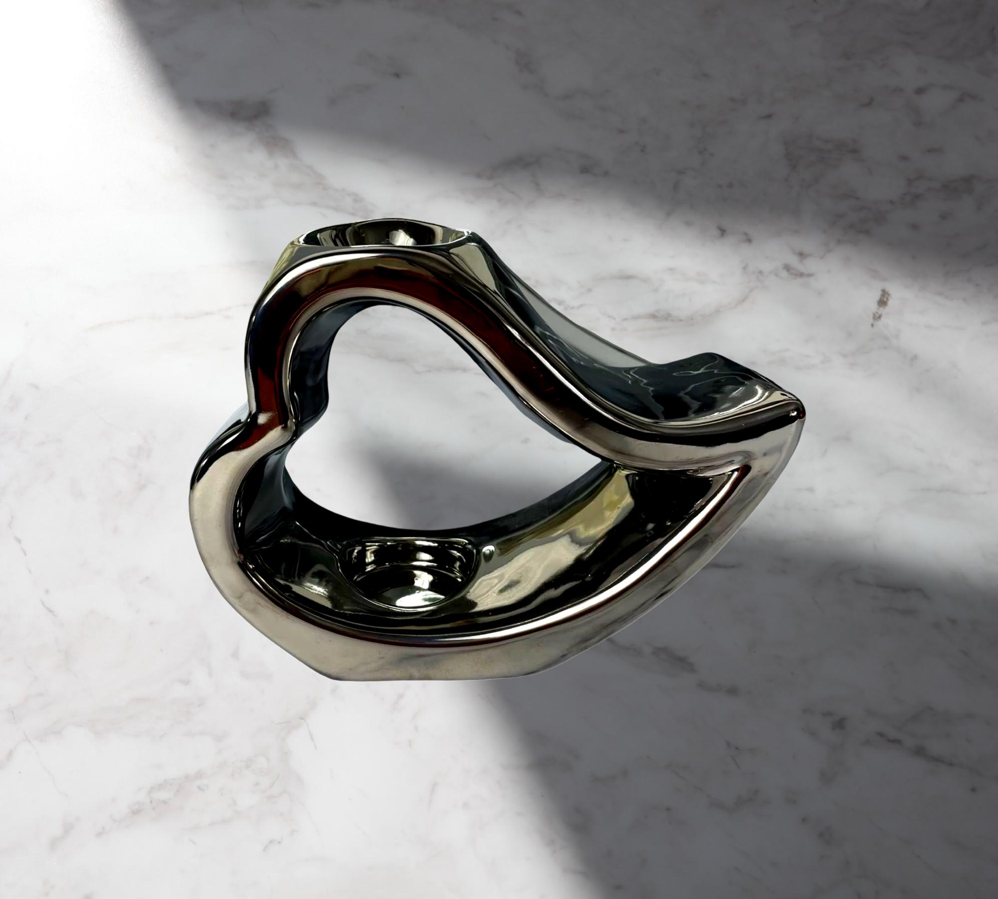 A metallic, abstract sculpture with a heart-like shape sits on a marble surface, reminiscent of The Soap Gal x's Silver Ceramic Sleepy Open Heart Romeo Oil Burner, casting intriguing shadows and evoking the allure of fragrance oils.