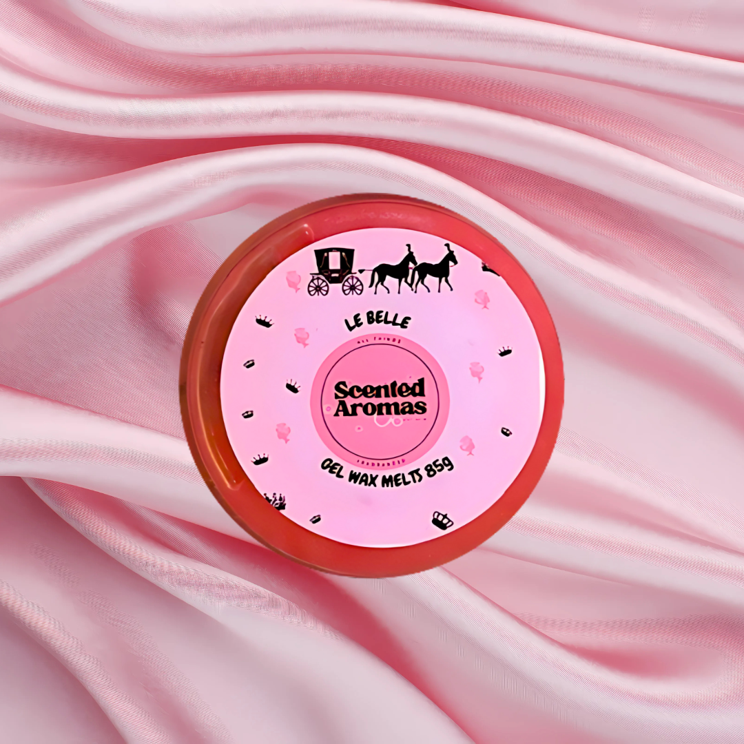 A round pink tin labeled "Le Est Belle Gel Wax Melts" by The Soap Gal x, featuring a charming carriage and horse design, rests on pink satin fabric. Inside, discover vegan wax melts that immerse you in luxurious fragrances, effortlessly blending elegance and sophistication.